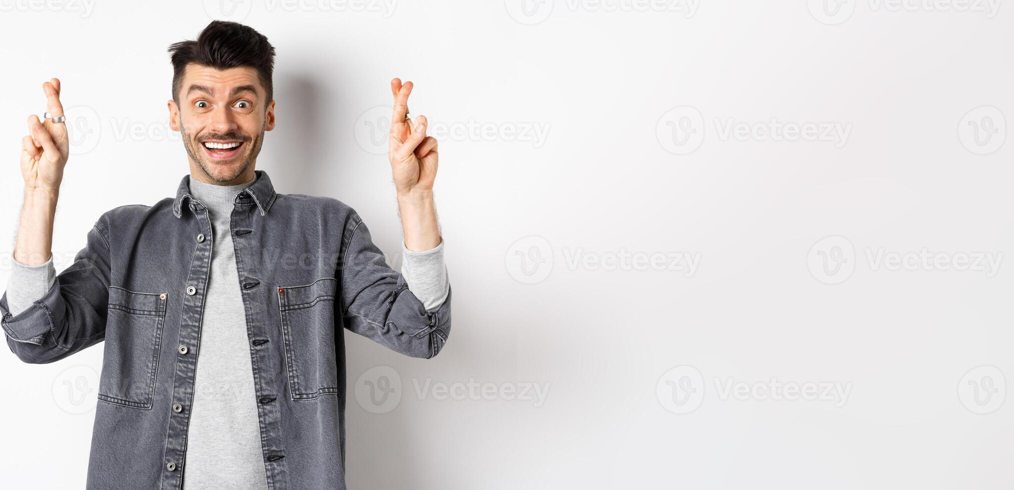 Young optimistic male hoping for dream come true, making wish with fingers crossed, smiling and expecting good news, standing on white background photo