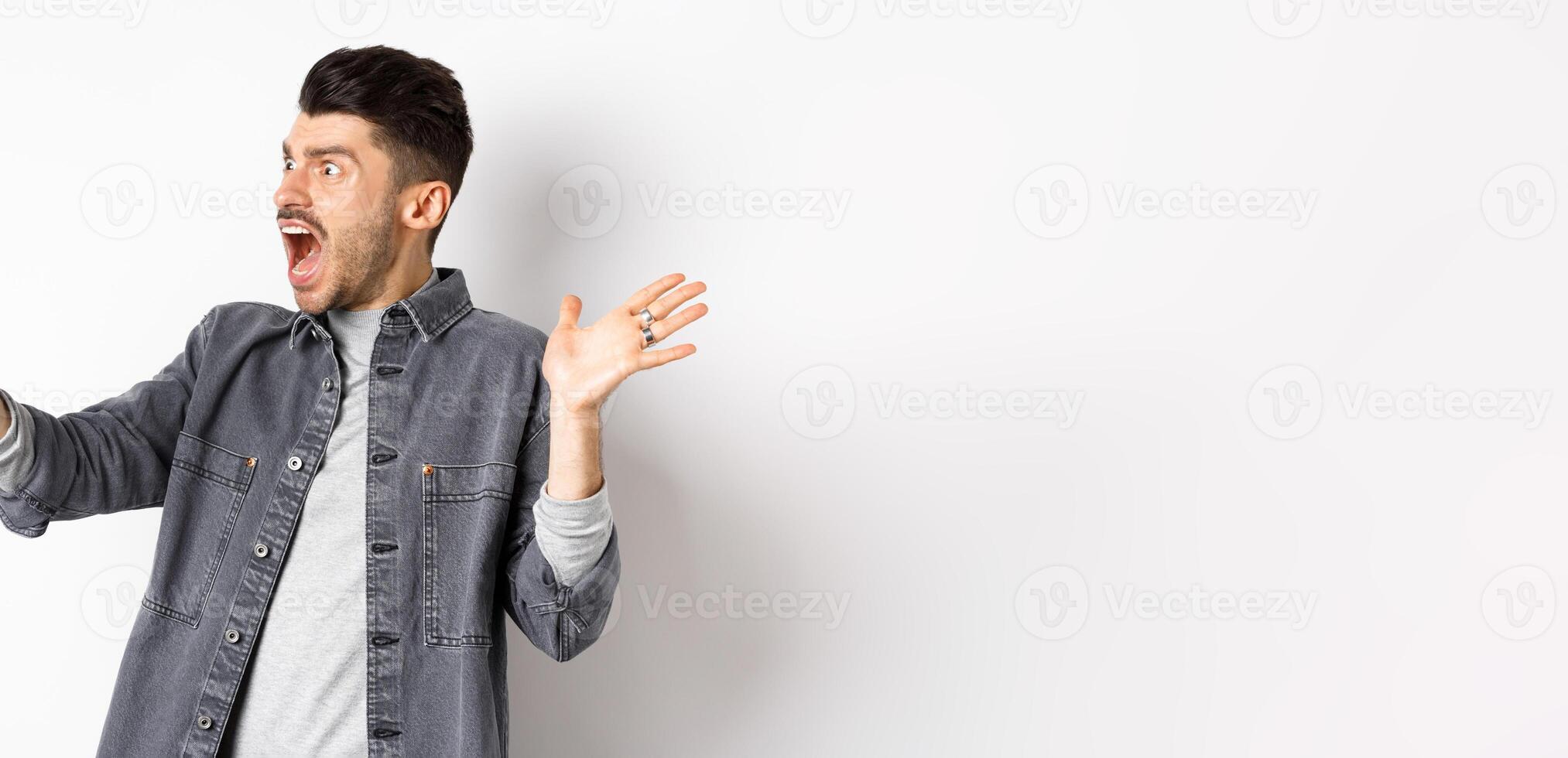 Shocked and confused man staring frustrated at smartphone screen, shouting from disappointment, standing against white background photo