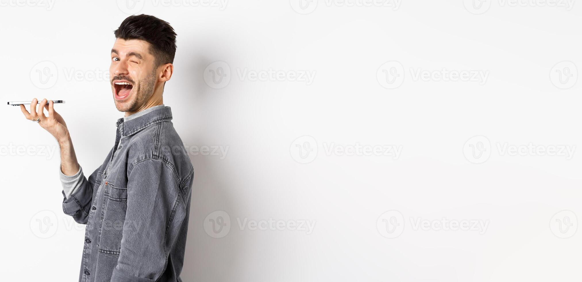 Happy guy winking at looking at camera while using voice translator app or talking on speakerphone, holding phone close to mouth, standing on white background photo