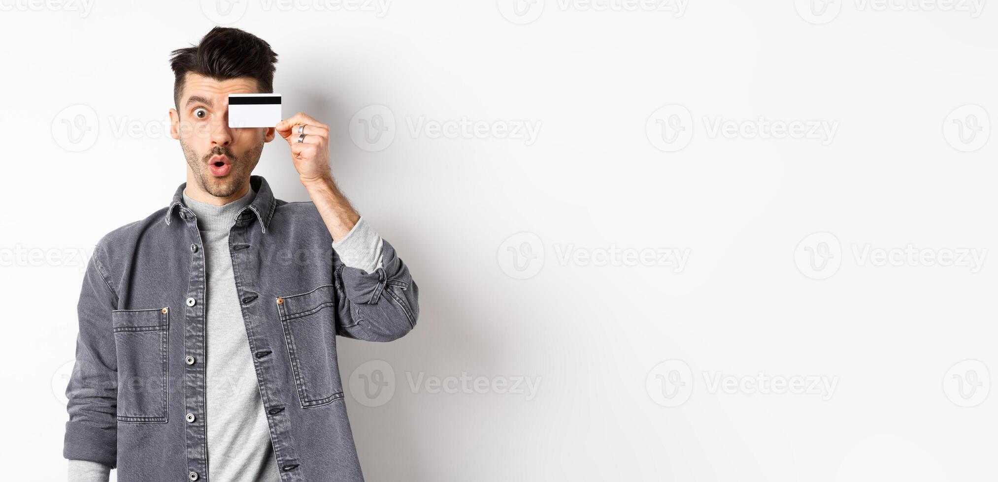 Excited funny man hold plastic credit card on eye and say wow, checking out special deal, standing on white background photo