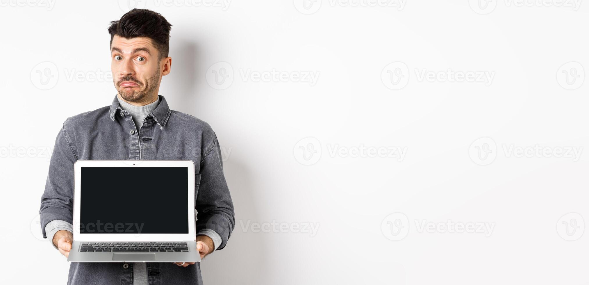 Indecisive young man pouting and shrugging while showing empty laptop screen, standing clueless on white background photo