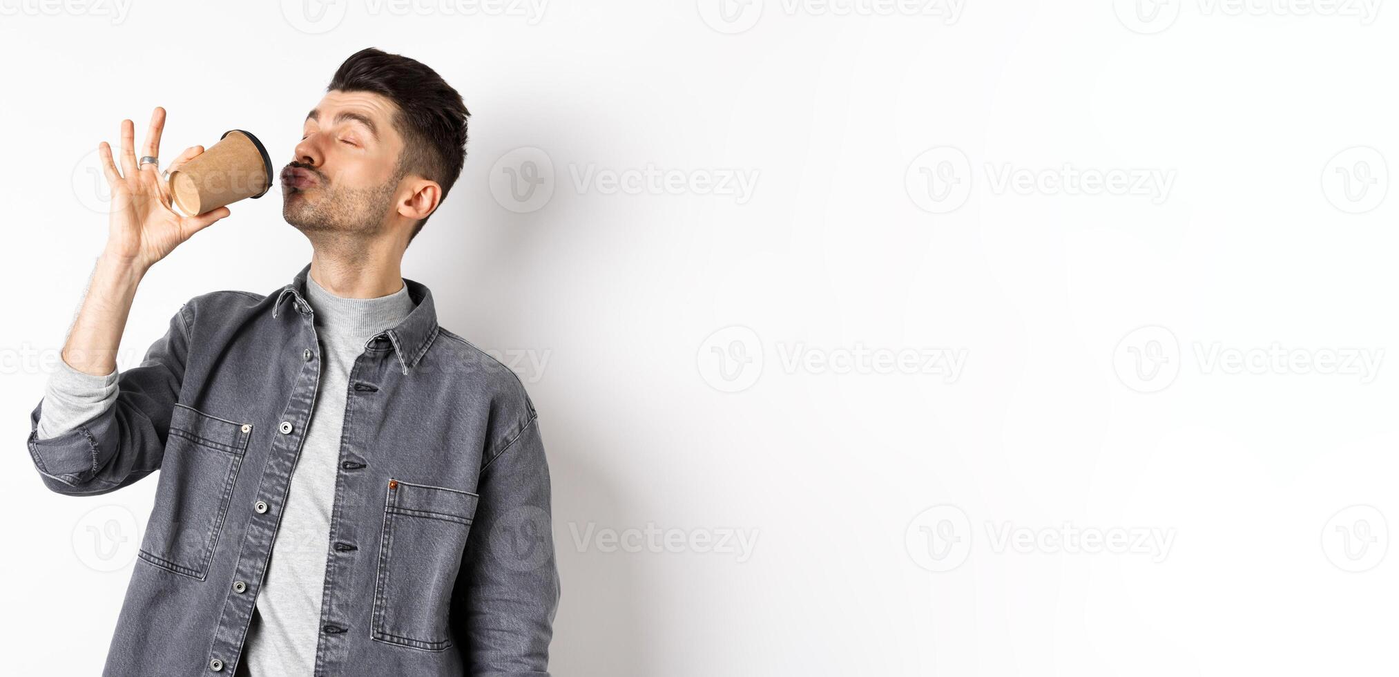 Funny guy enjoying drinking coffee from cup, sipping takeaway drink from cafe with satisfaction, standing on white background photo