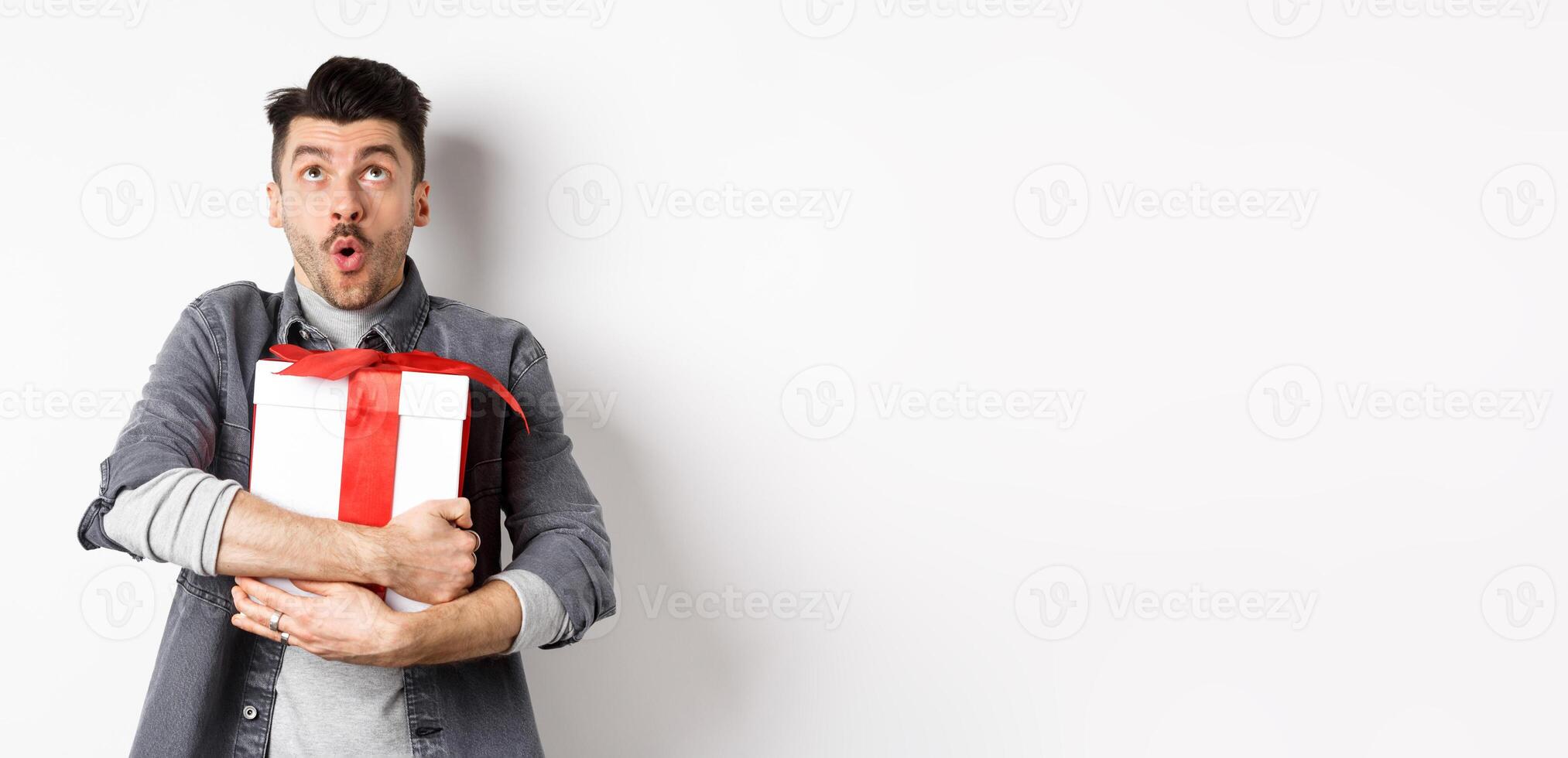Surprised funny guy hugging big holiday gift and looking up, say wow amazed, checking out Valentines day special offer, standing on white background photo