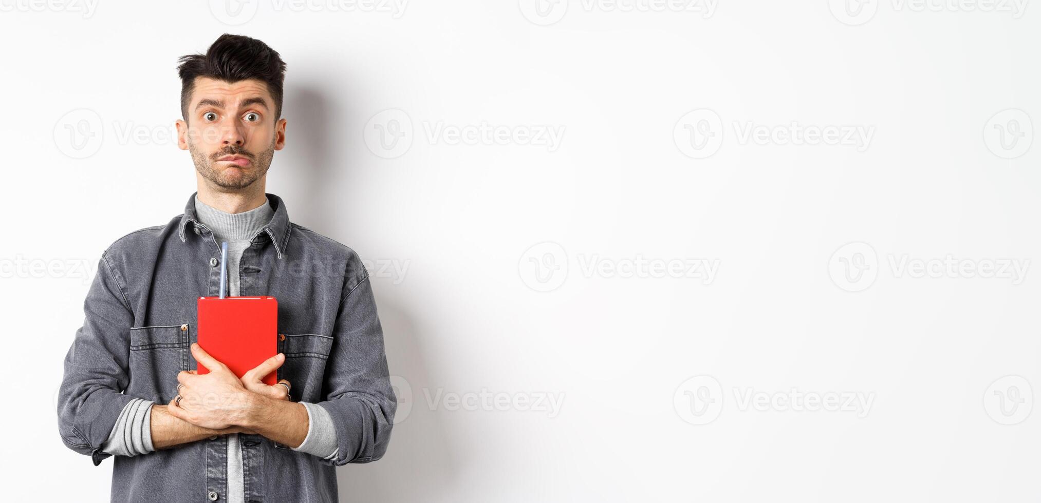 Cute and shy guy hugging his diary, holding red journal and look modest at camera, standing against white background photo