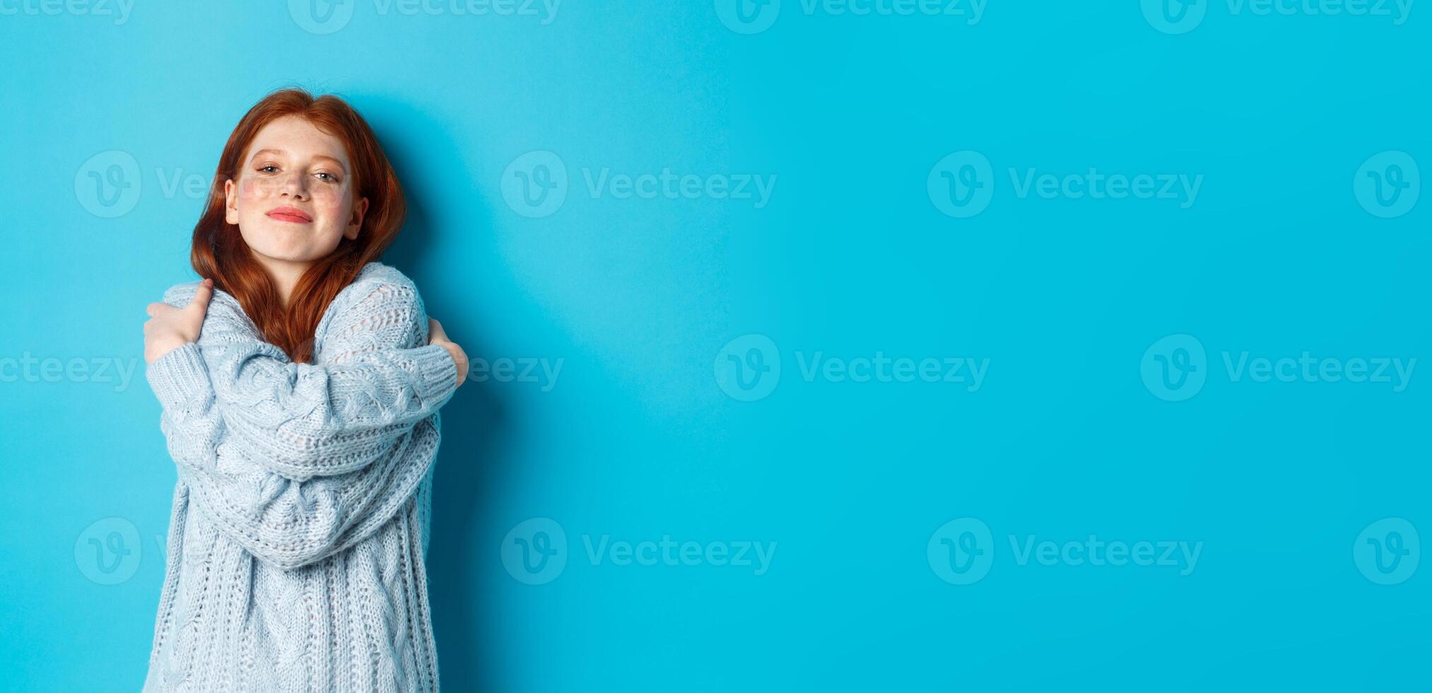 Happy cute redhead girl hugging herself, wearing comfortable and warm sweater, smiling at camera, standing over blue background photo