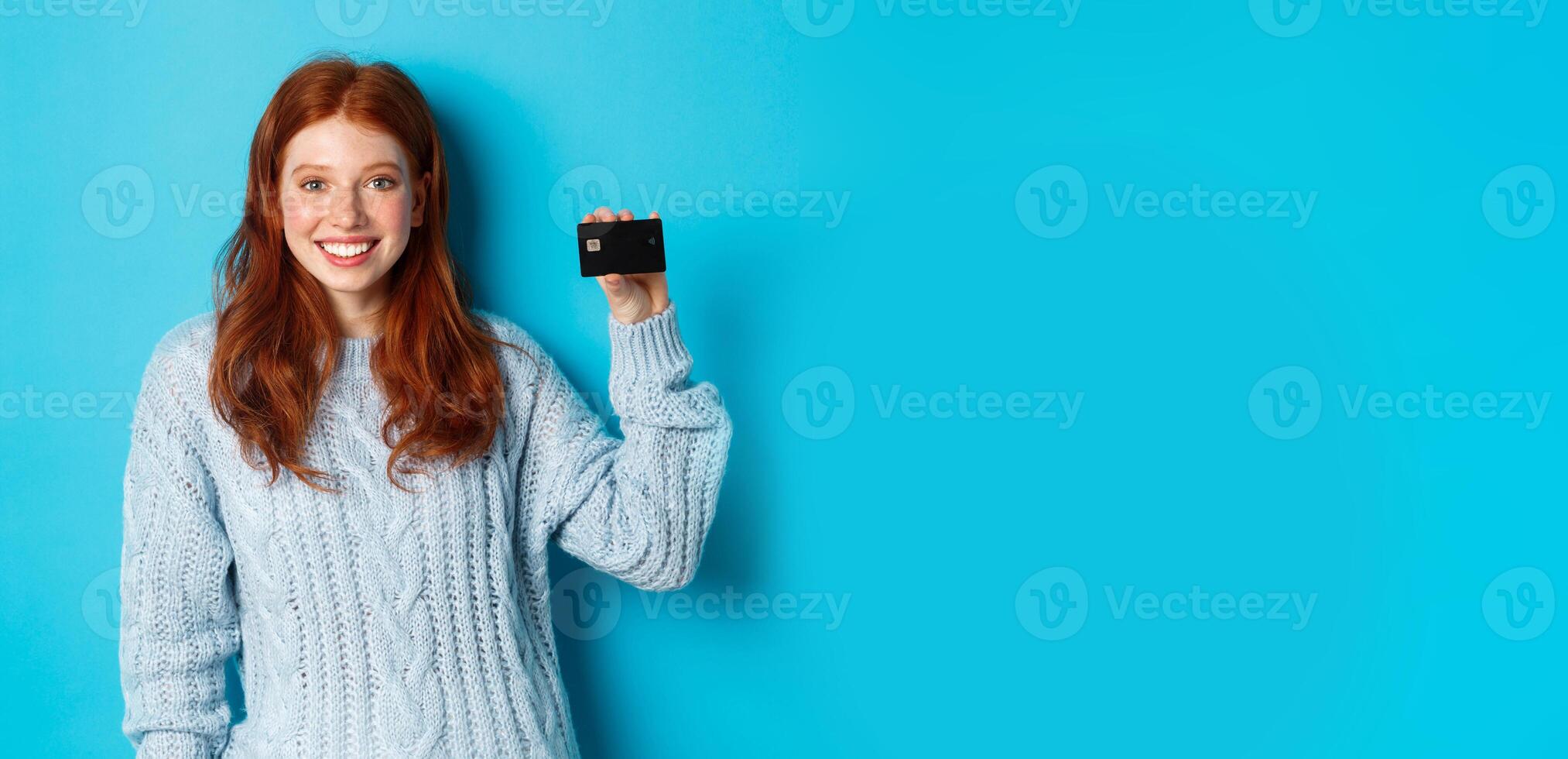 Cute redhead girl in sweater showing credit card, smiling at camera, standing over blue background photo
