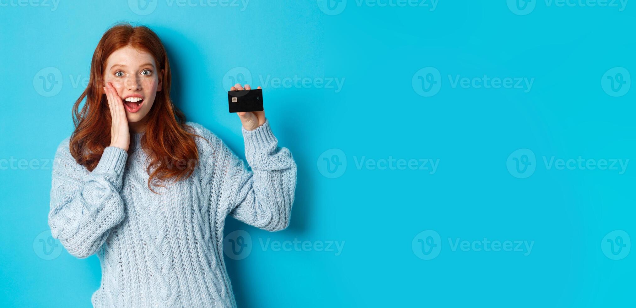 Cute redhead girl in sweater showing credit card, smiling at camera, standing over blue background photo