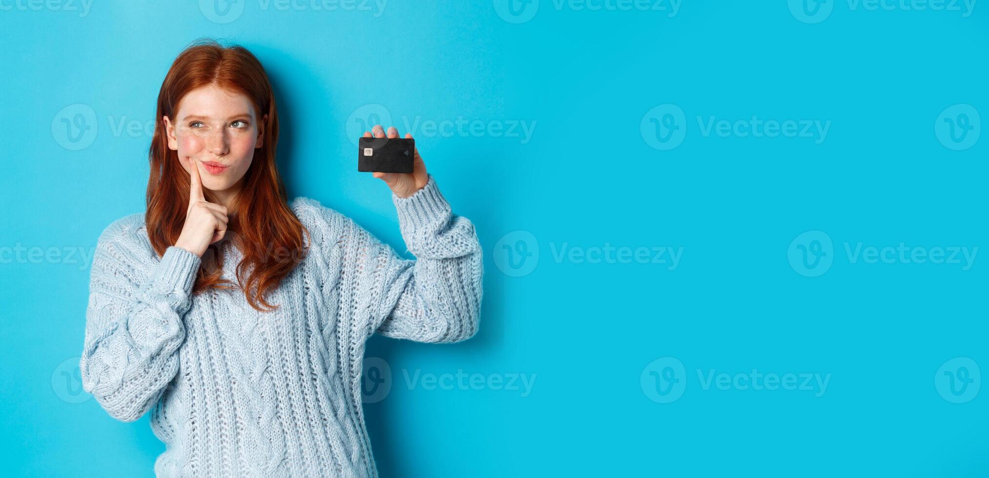 Image of thoughtful redhead girl thinking about shopping, showing credit card and pondering, standing over blue background photo