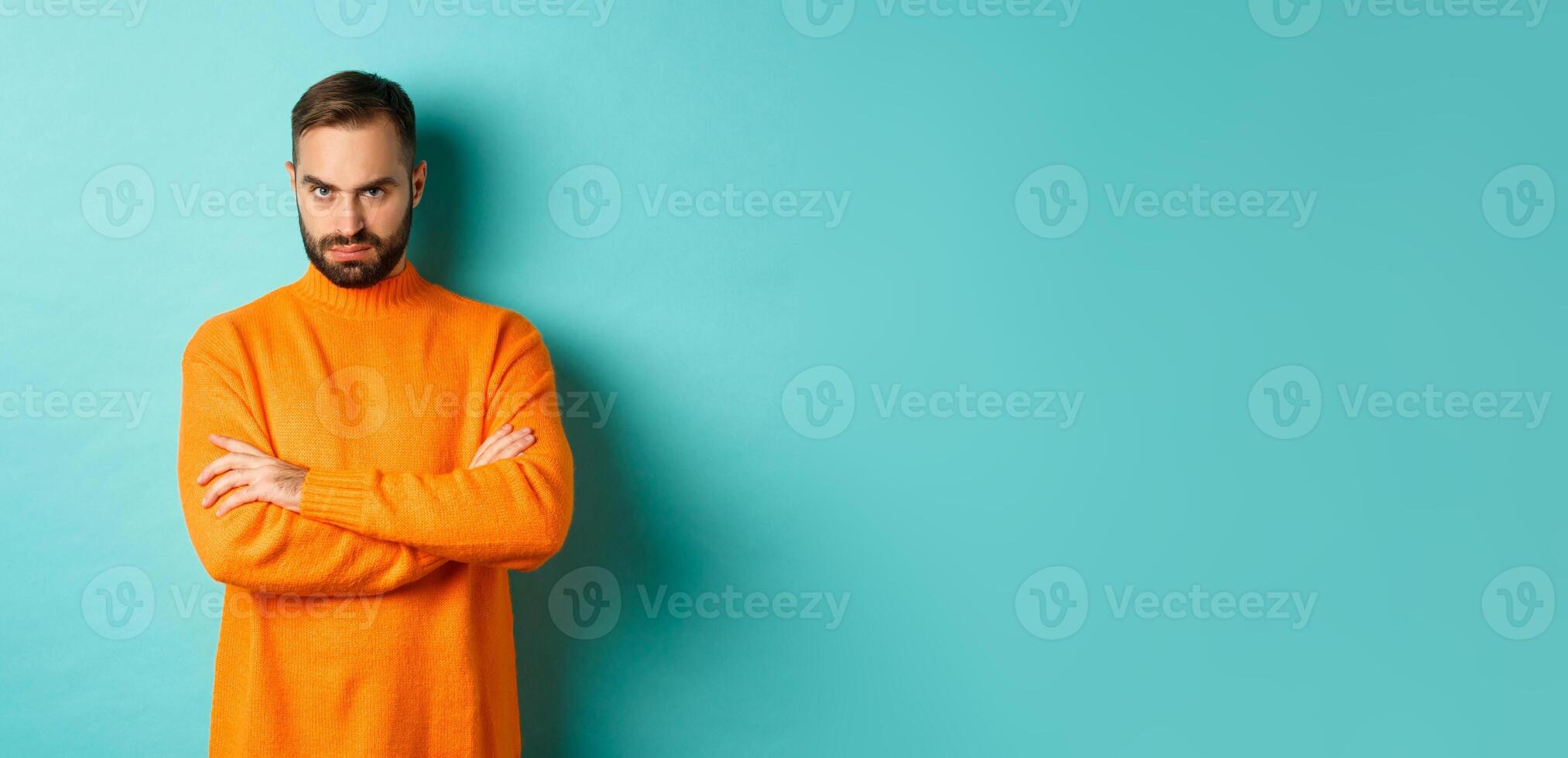 Offended man looking angry at you, cross arms on chest and stare mad, standing in orange sweater against turquoise background photo