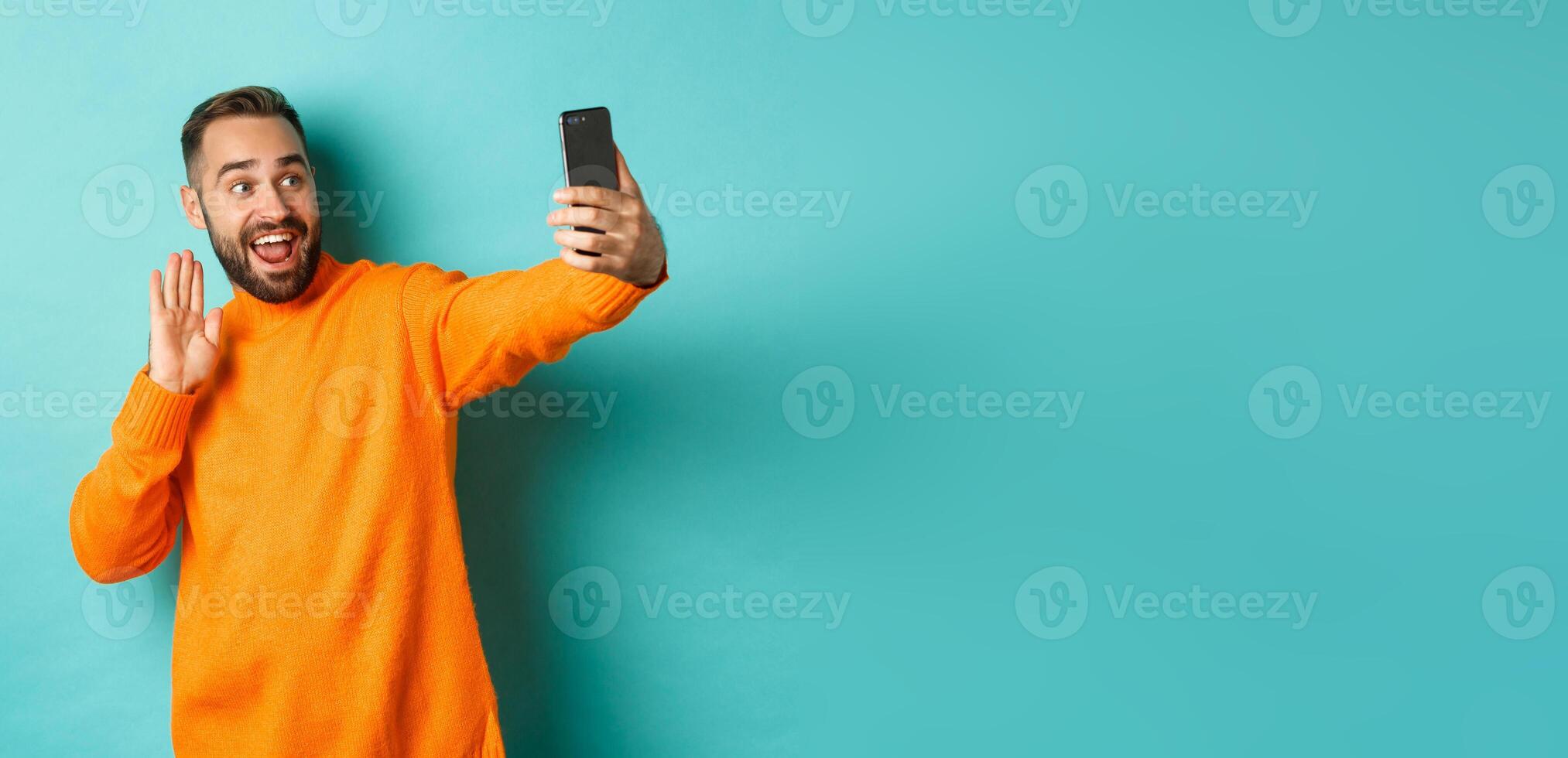 Happy young man calling, talking online with mobile phone, saying hello to smartphone camera and waving hand friendly, standing over light blue background photo
