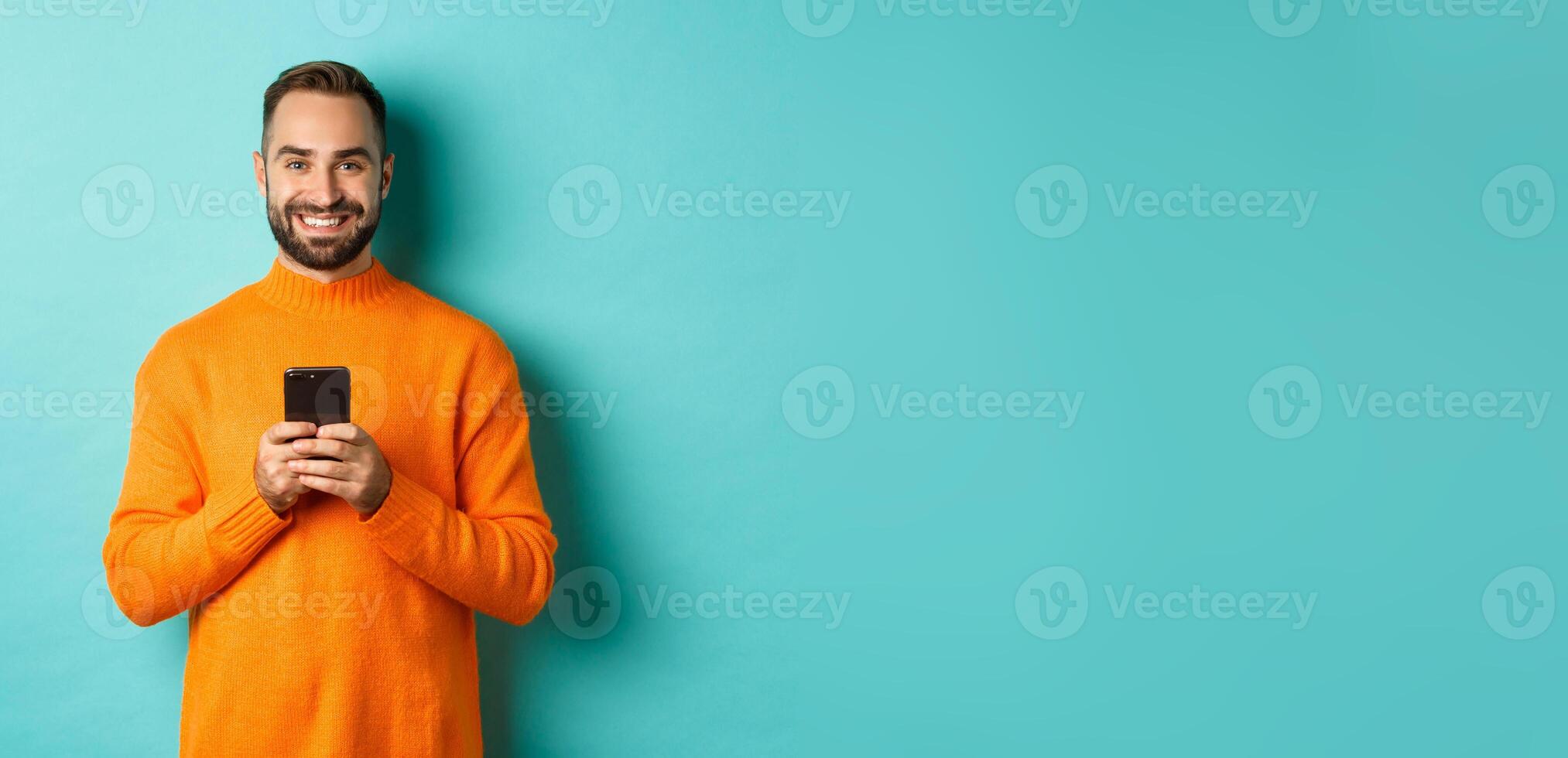 Happy handsome man writing message on mobile phone, holding smartphone and smiling, standing against turquoise background photo