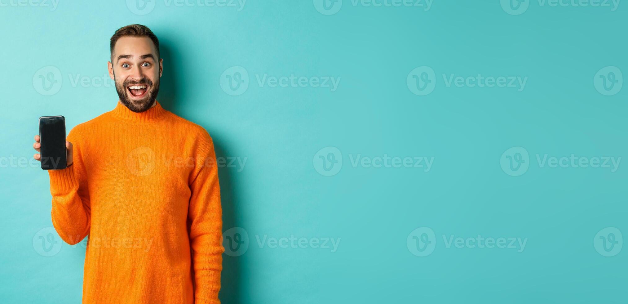 Photo of happy man showing mobile screen, introduce online store, application, standing over turquoise background