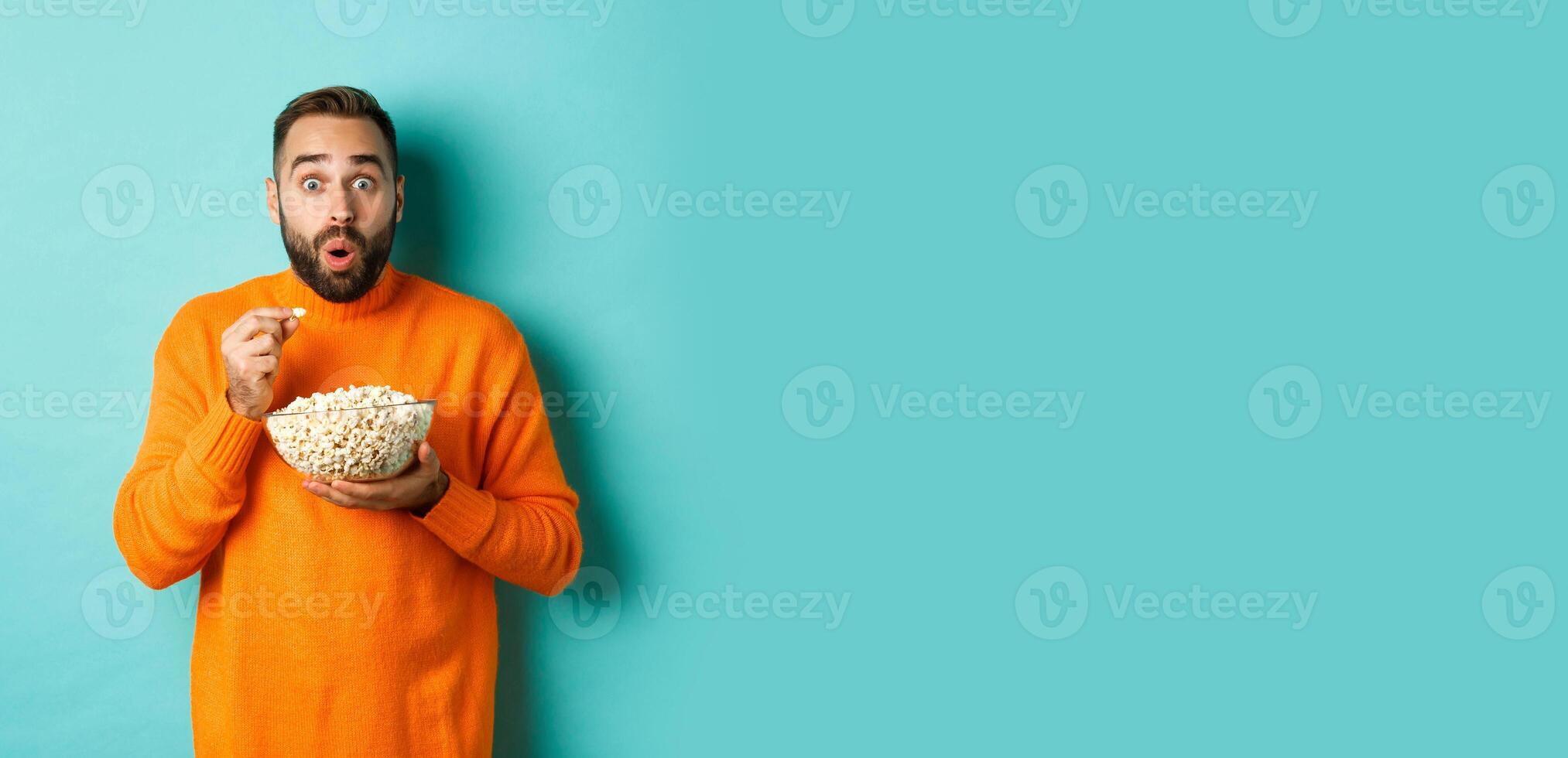 Excited young man watching interesting movie on tv screen, eating popcorn and looking amazed, blue background photo