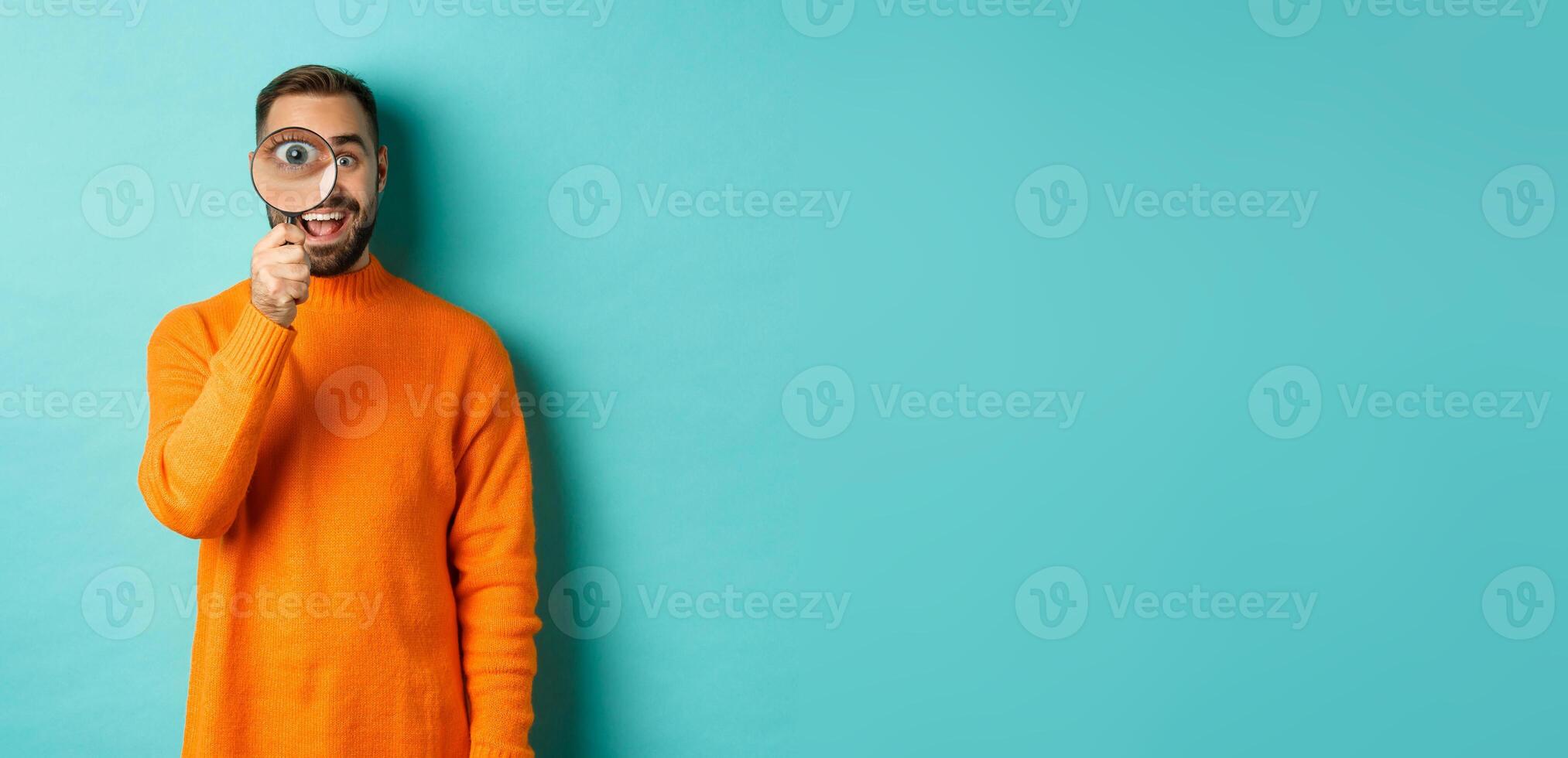 Cheerful man searching for something, looking through magnifying glass and smiling happy, standing over blue background photo