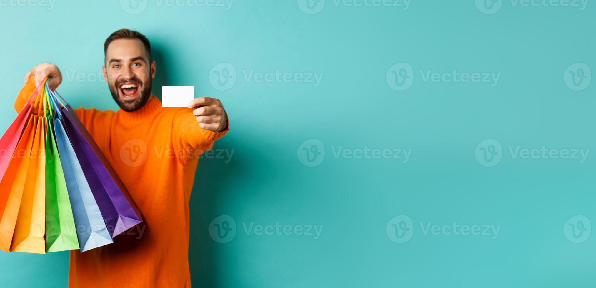Happy aduly man showing credit card and shopping bags, standing against turquoise background photo