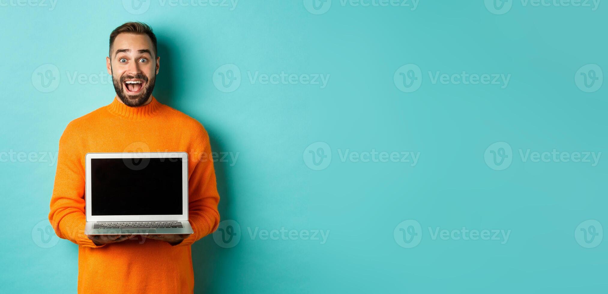 Handsome bearded man in orange sweater showing laptop screen, demonstrating promo, standing over light blue background photo