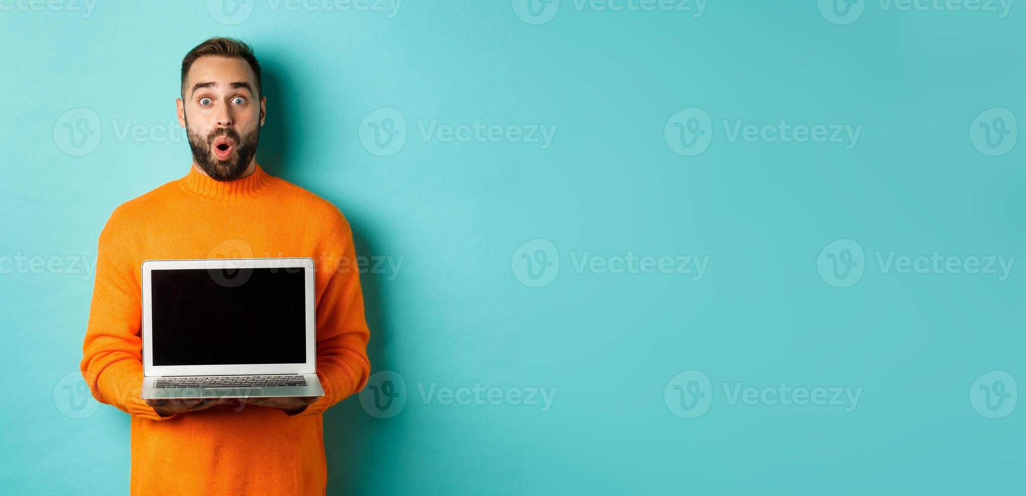 Handsome bearded man in orange sweater showing laptop screen, demonstrating promo, standing over light blue background photo