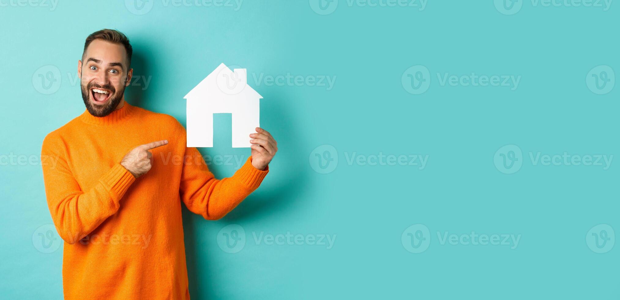 Real estate concept. Happy man pointing finger at paper home maket and smiling, standing over light blue background photo