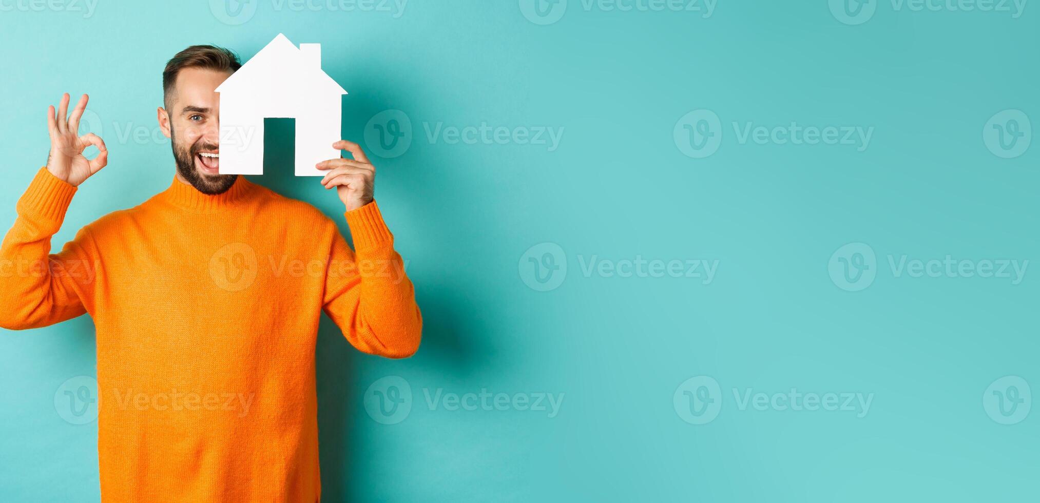 Real estate concept. Satisfied man recommending agency, showing okay sign and house maket, standing over blue background photo