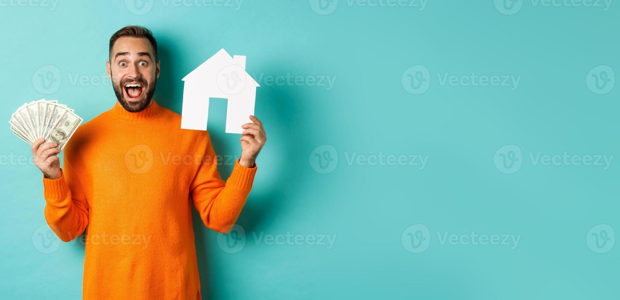 Real estate concept and mortgage concept. Excited man showing dollars and paper maket of house, buying flat, standing over blue background photo