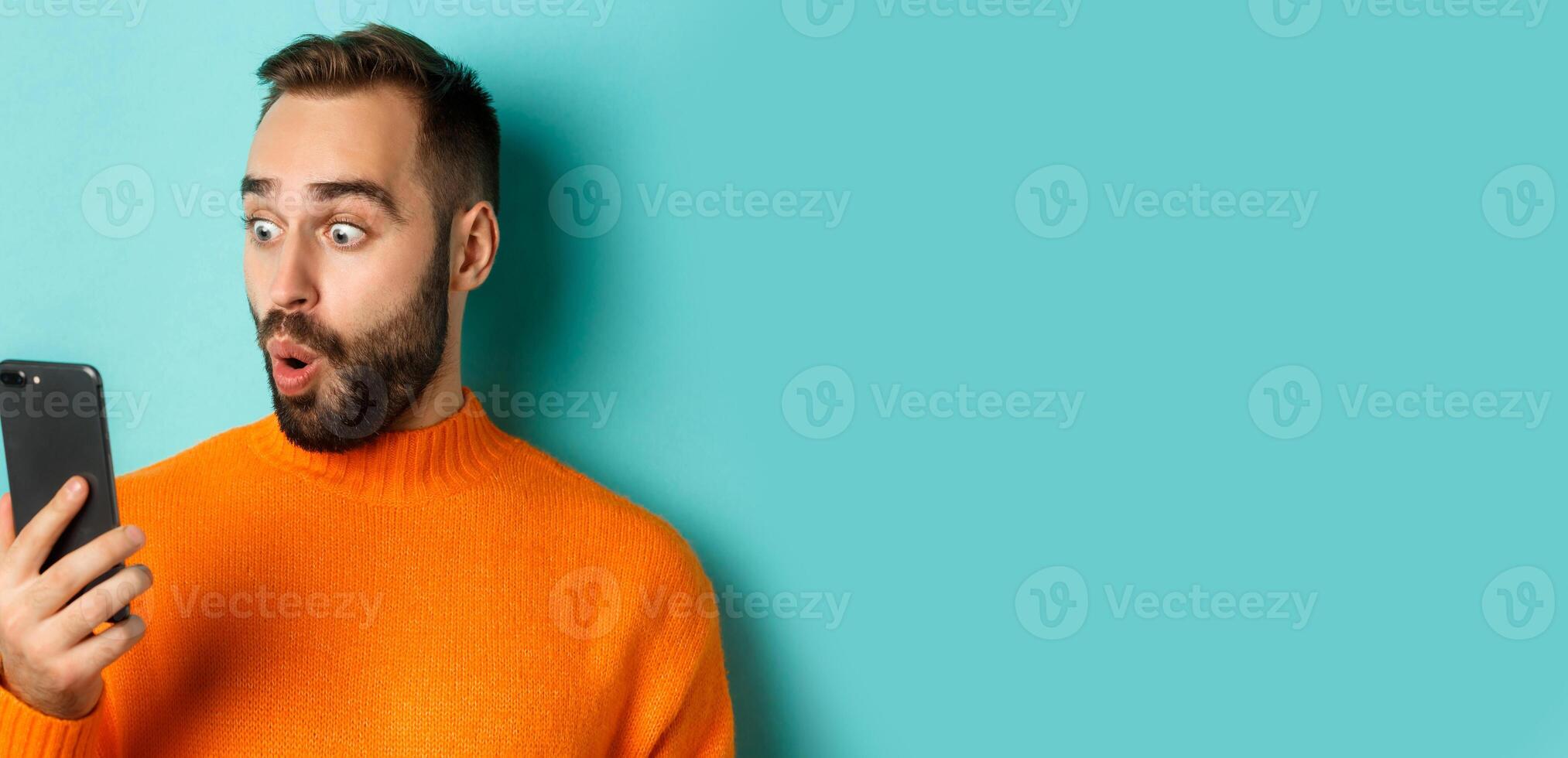 Close-up of caucasian man staring at phone screen with surprised face, wearing orange sweater, light blue background photo