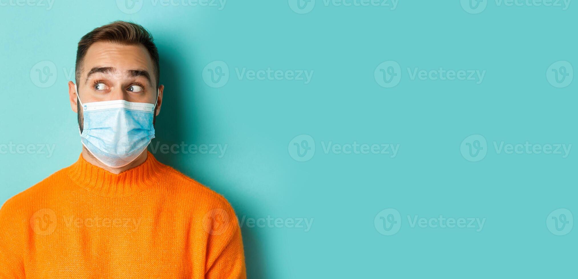 Covid-19, social distancing and quarantine concept. Close-up of young male model in medical mask looking left curious, staring at promo copy space, light blue background photo