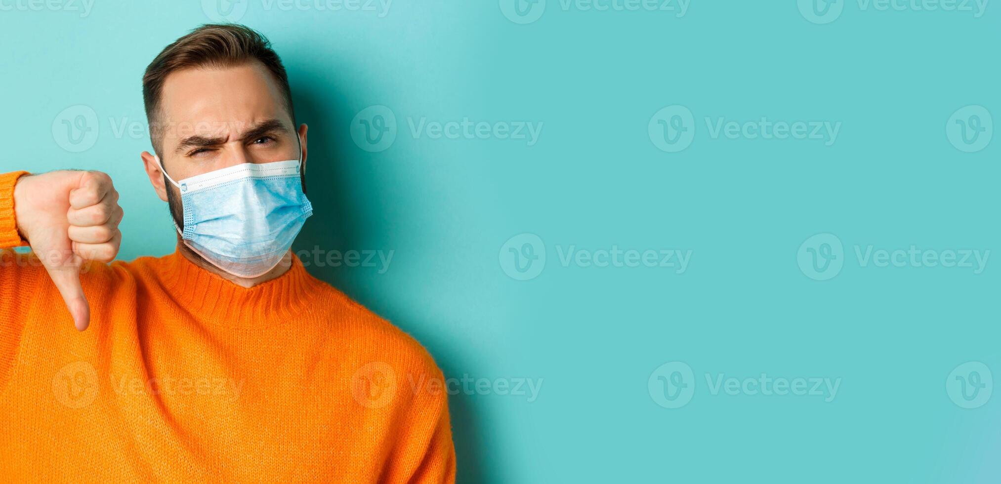 Covid-19, social distancing and quarantine concept. Close-up of disappointed male model in medical mask, showing thumb-down, negative feedback, standing over light blue background photo