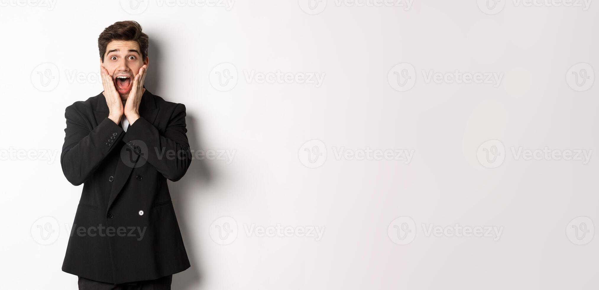 Portrait of surprised handsome businessman in suit, reacting to holidays promo, looking amazed at camera, standing over white background photo