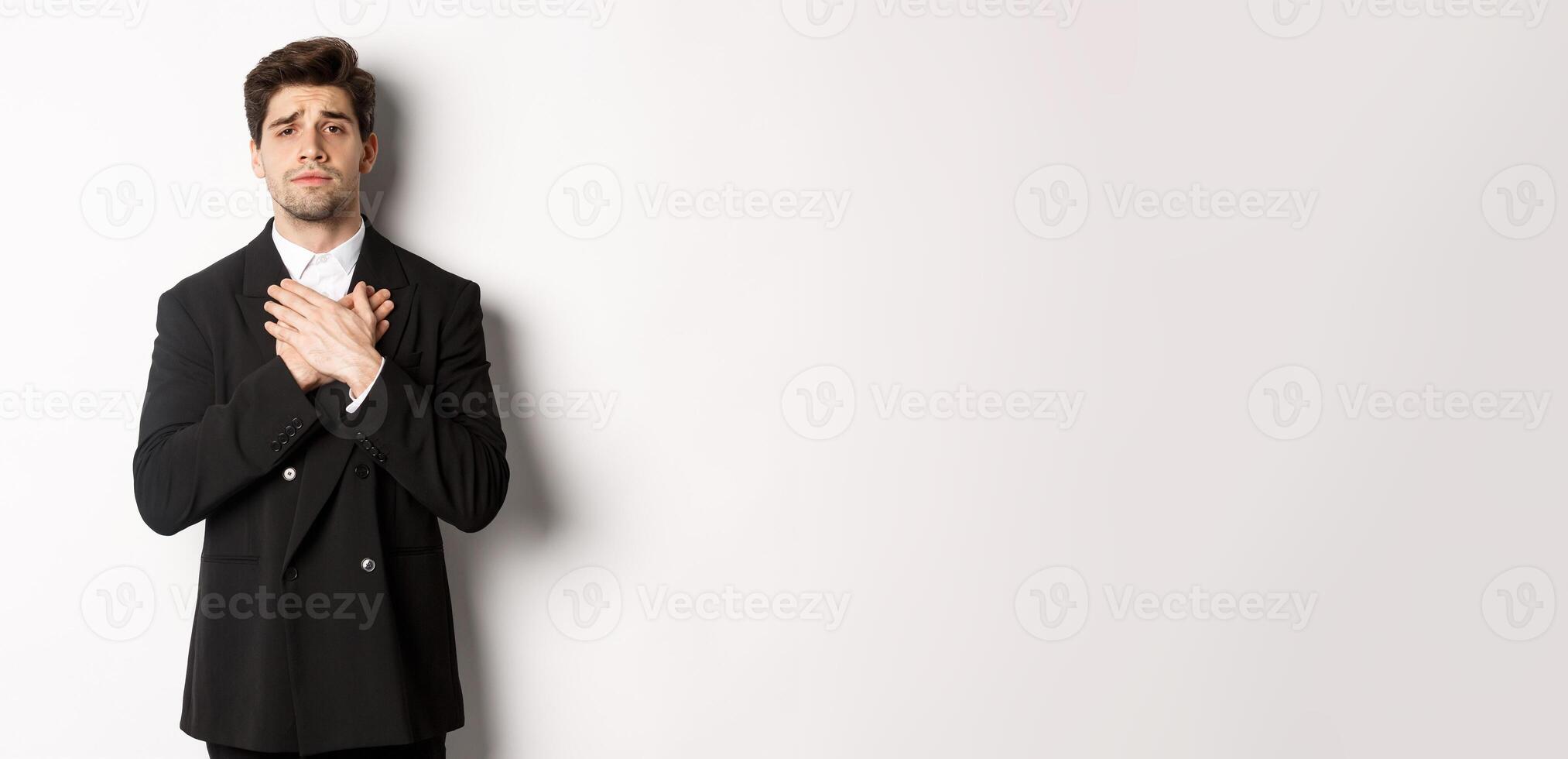 Portrait of touched and compassionate guy in suit, holding hands on heart and looking with pity at camera, standing over white background photo