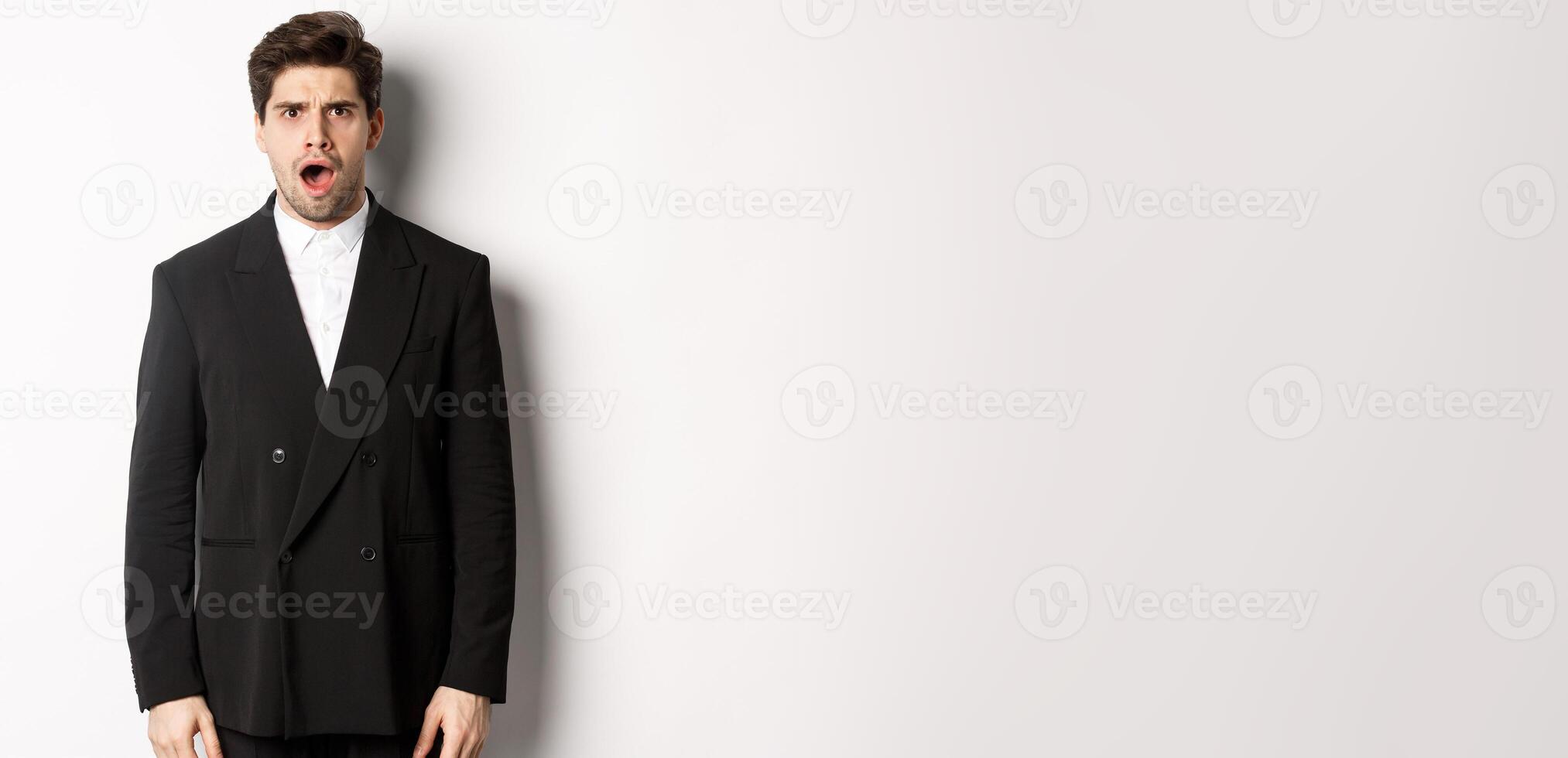 Portrait of shocked and startled handsome man in suit, drop jaw and looking in awe at camera, standing over white background photo