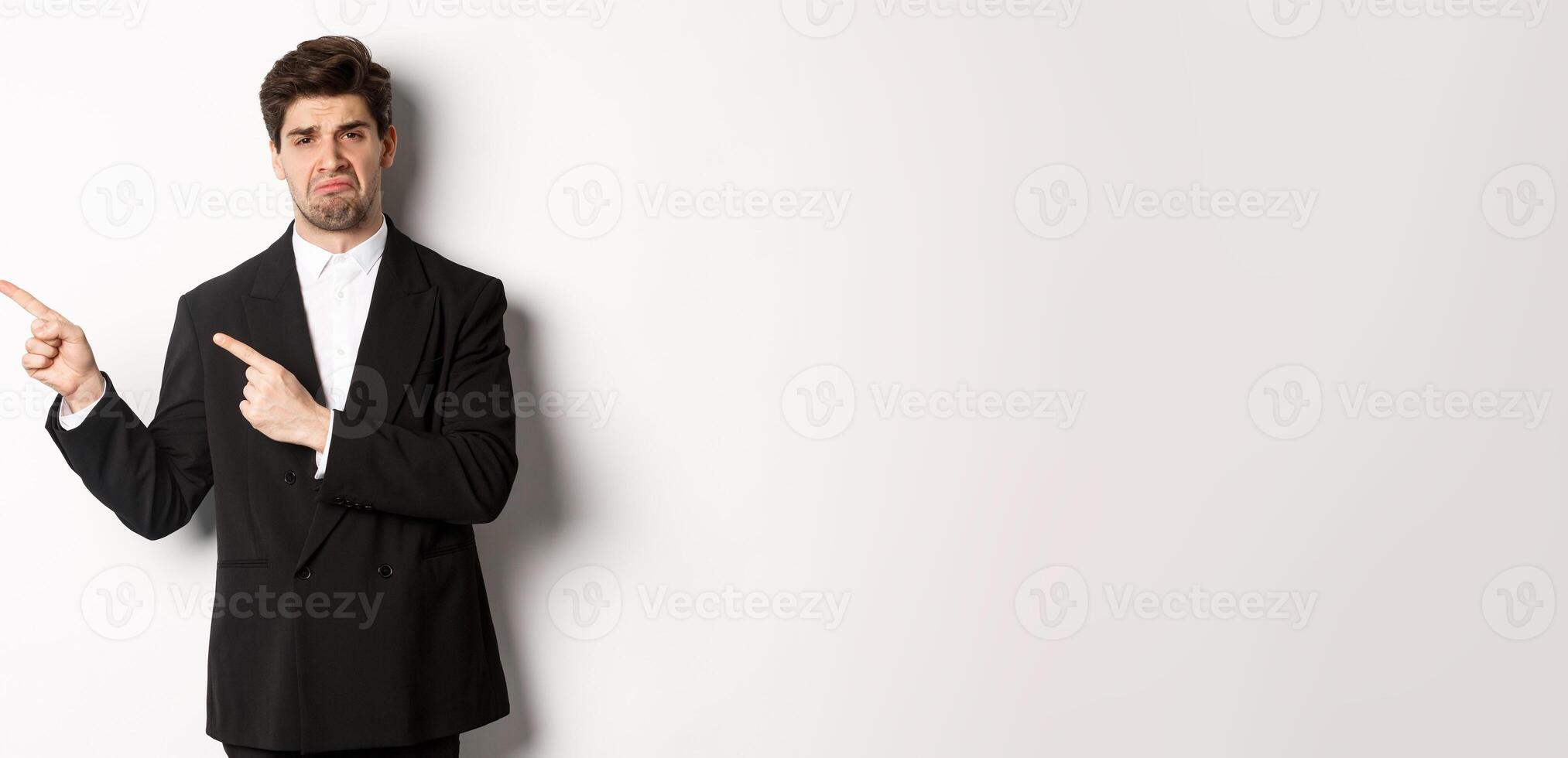 Portrait of disappointed and sad handsome businessman in suit, complaining and pointing fingers left at something bad, standing upset against white background photo