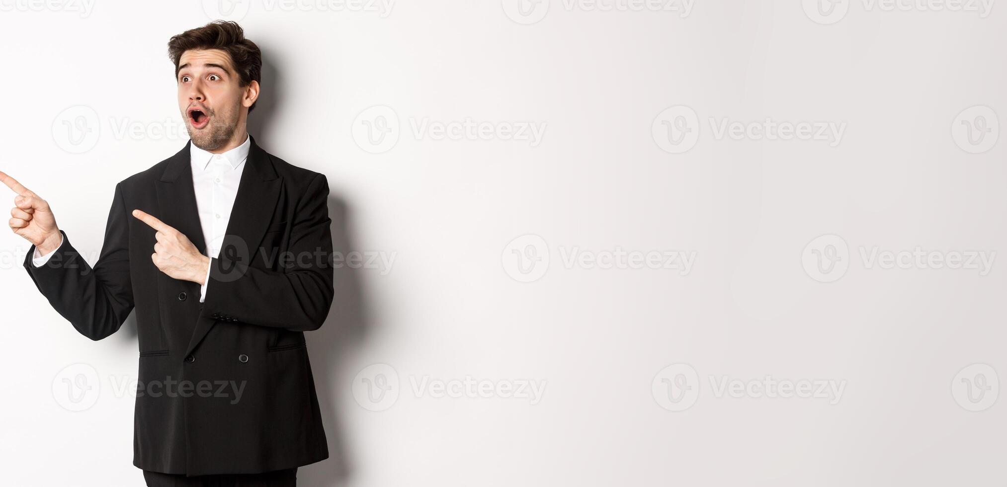 Impressed handsome man in party suit, looking at new year promo offer and pointing fingers left at banner, standing over white background photo
