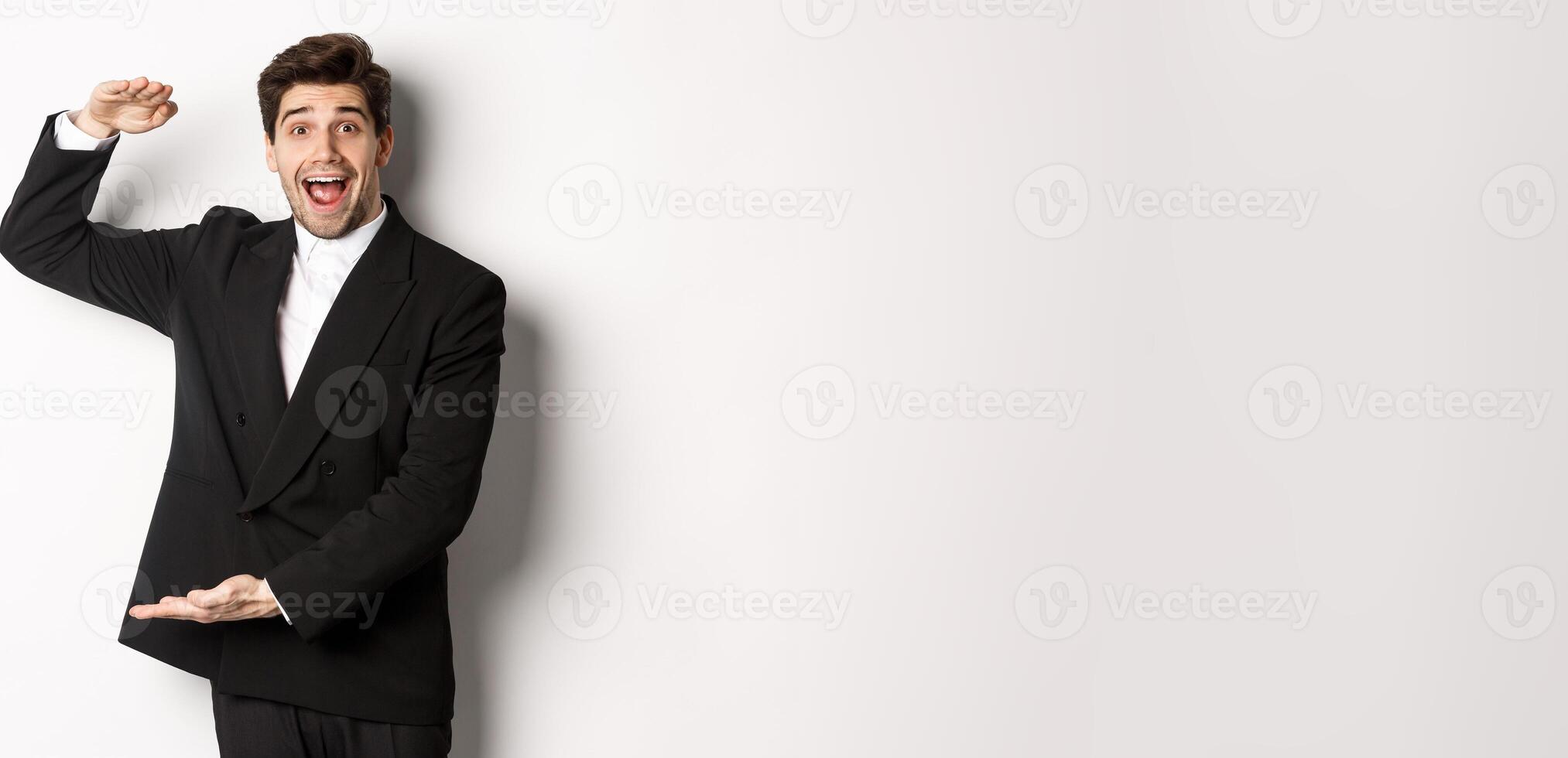 Portrait of excited handsome man in suit, shaping big object on copy space and smiling amazed, holding something, standing over white background photo