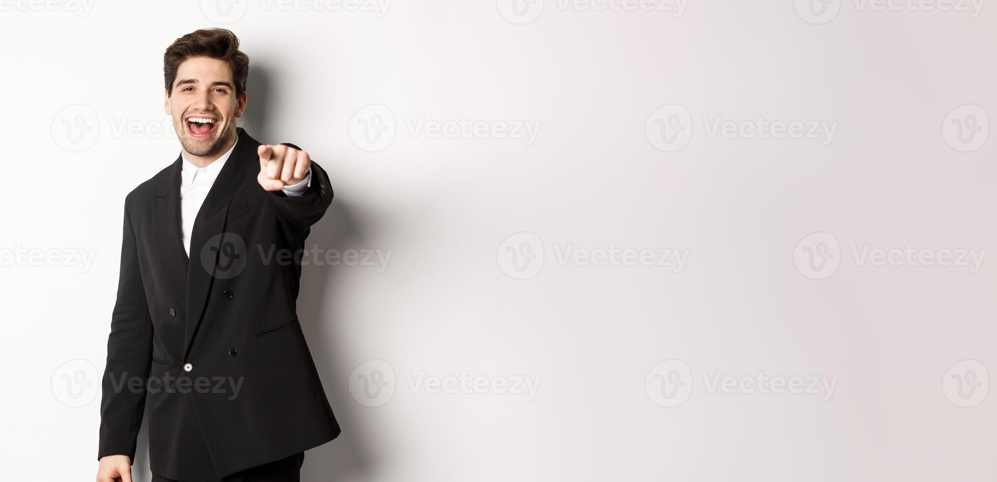Concept of new year party, celebration and lifestyle. Portrait of handsome stylish man in black suit, smiling and pointing finger at camera, standing over white background photo