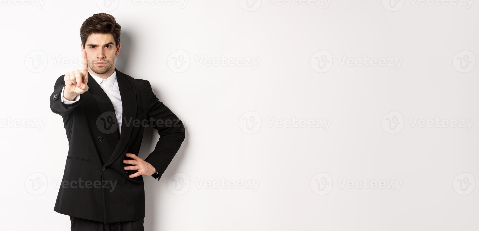 Portrait of serious handsome man in business suit, showing one finger to prohibit or decline something, telling to stop, disagree with you, standing over white background photo