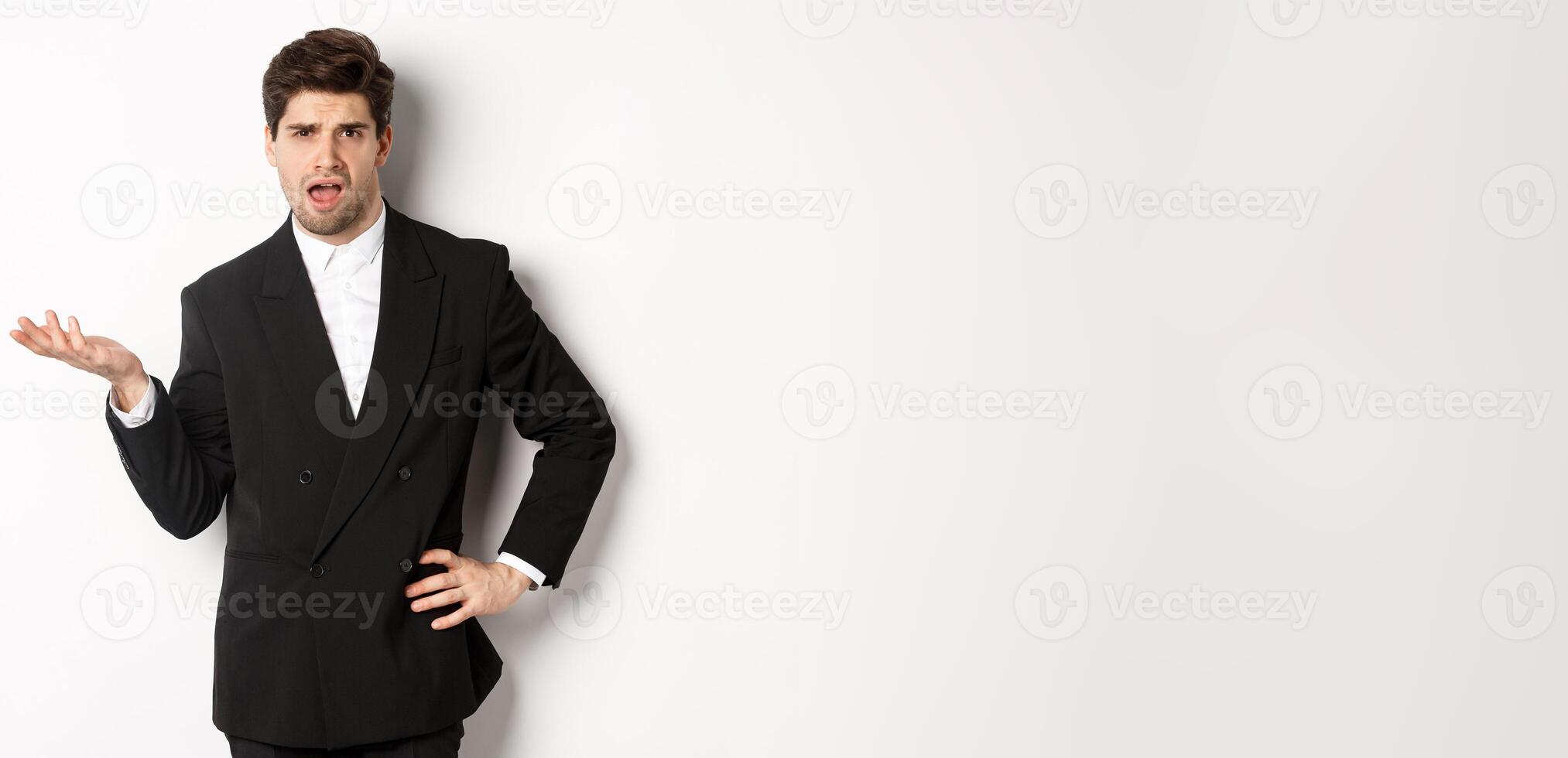 Portrait of arrogant man in black suit, looking confused and disappointed, complaining about something strange, standing over white background photo