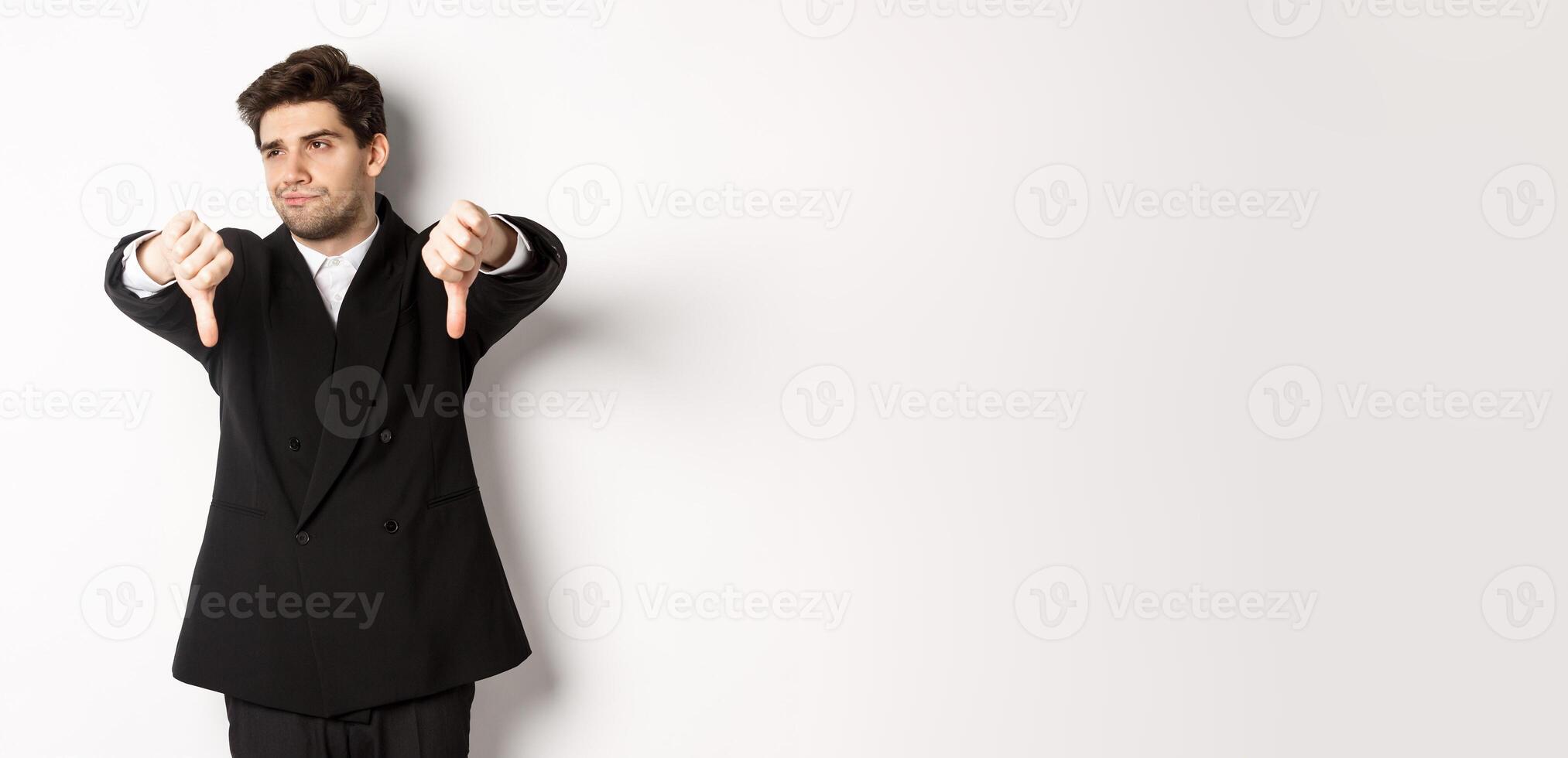 Skeptical and displeased man in black suit, looking away reluctant, showing thumbs-down, dislike something, standing over white background photo