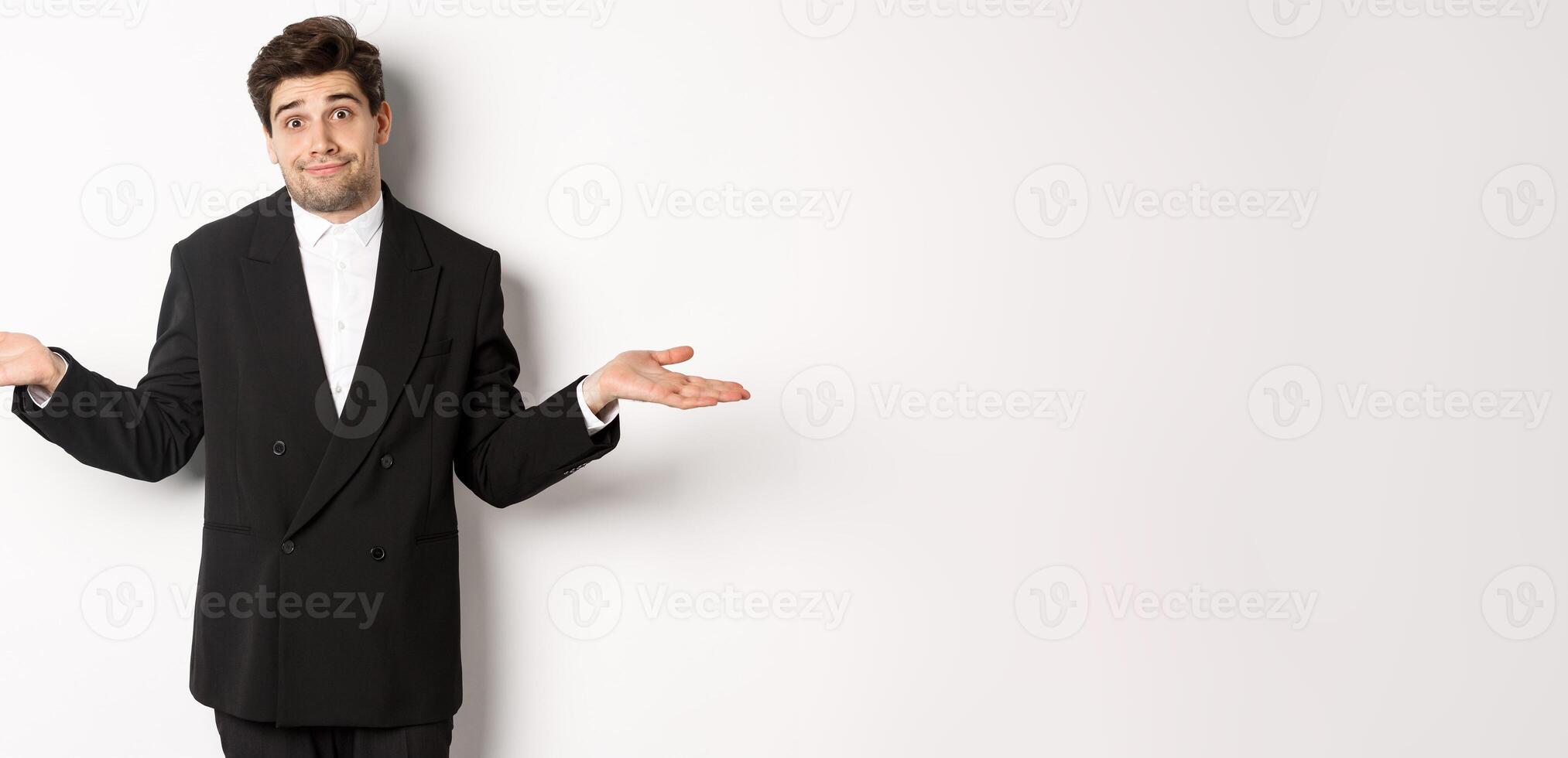Image of handsome confused man in black suit, shrugging and looking clueless, dont know anything, standing over white background photo