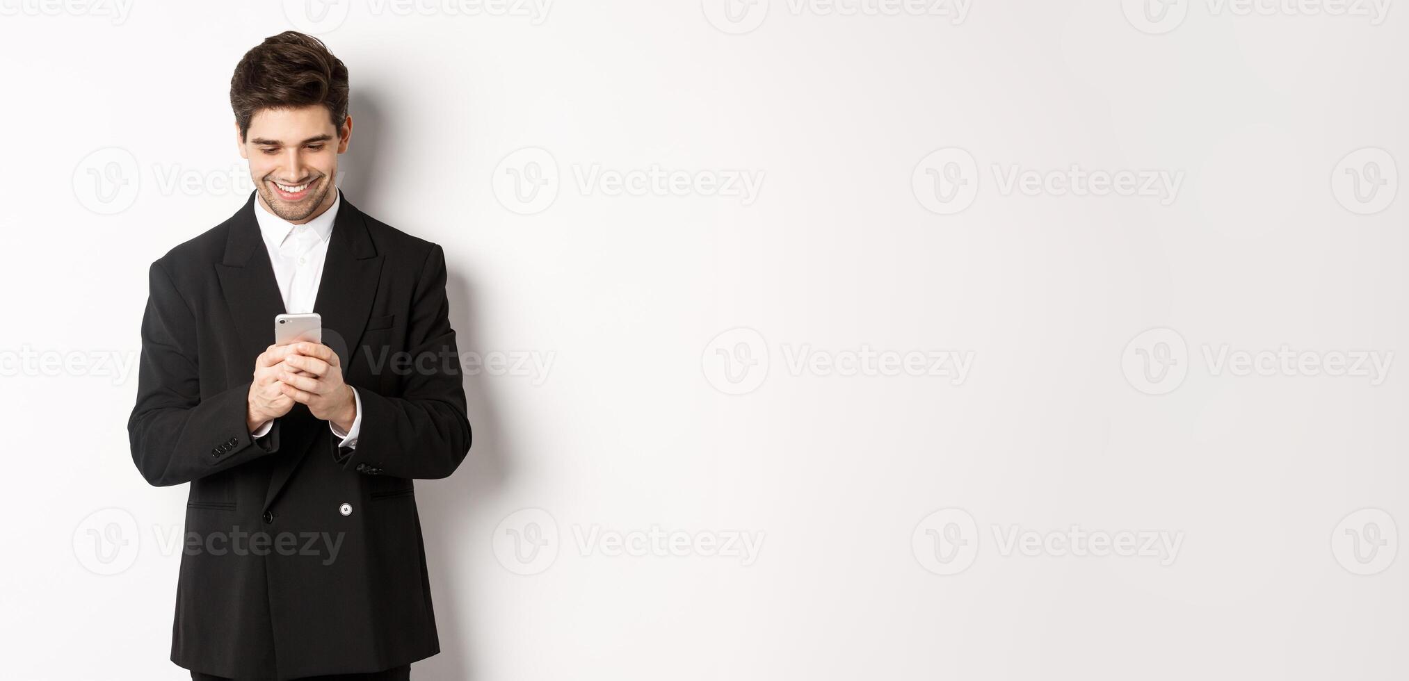 Portrait of handsome stylish businessman in black suit, writing a message, smiling and looking at smartphone, standing over white background photo