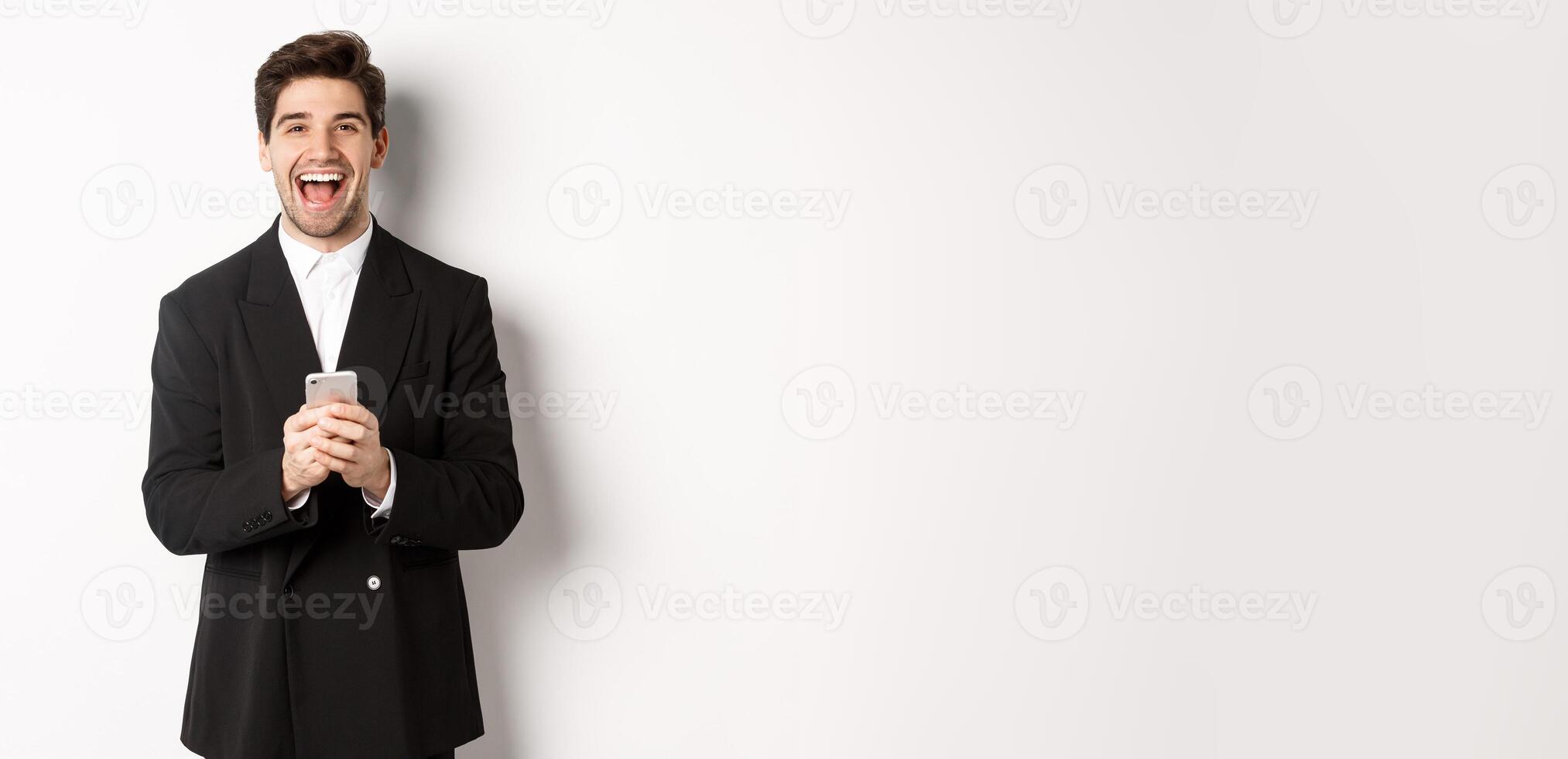 Portrait of happy good-looking man, wearing black suit, laughing from happiness and using mobile phone, standing over white background photo