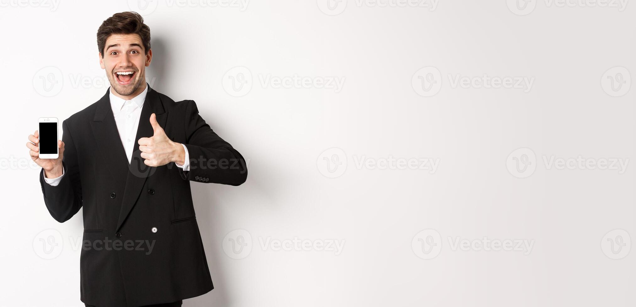 Image of handsome male entrepreneur in black suit, recommending app or online shop, showing thumbs-up and smartphone screen, standing over white background photo