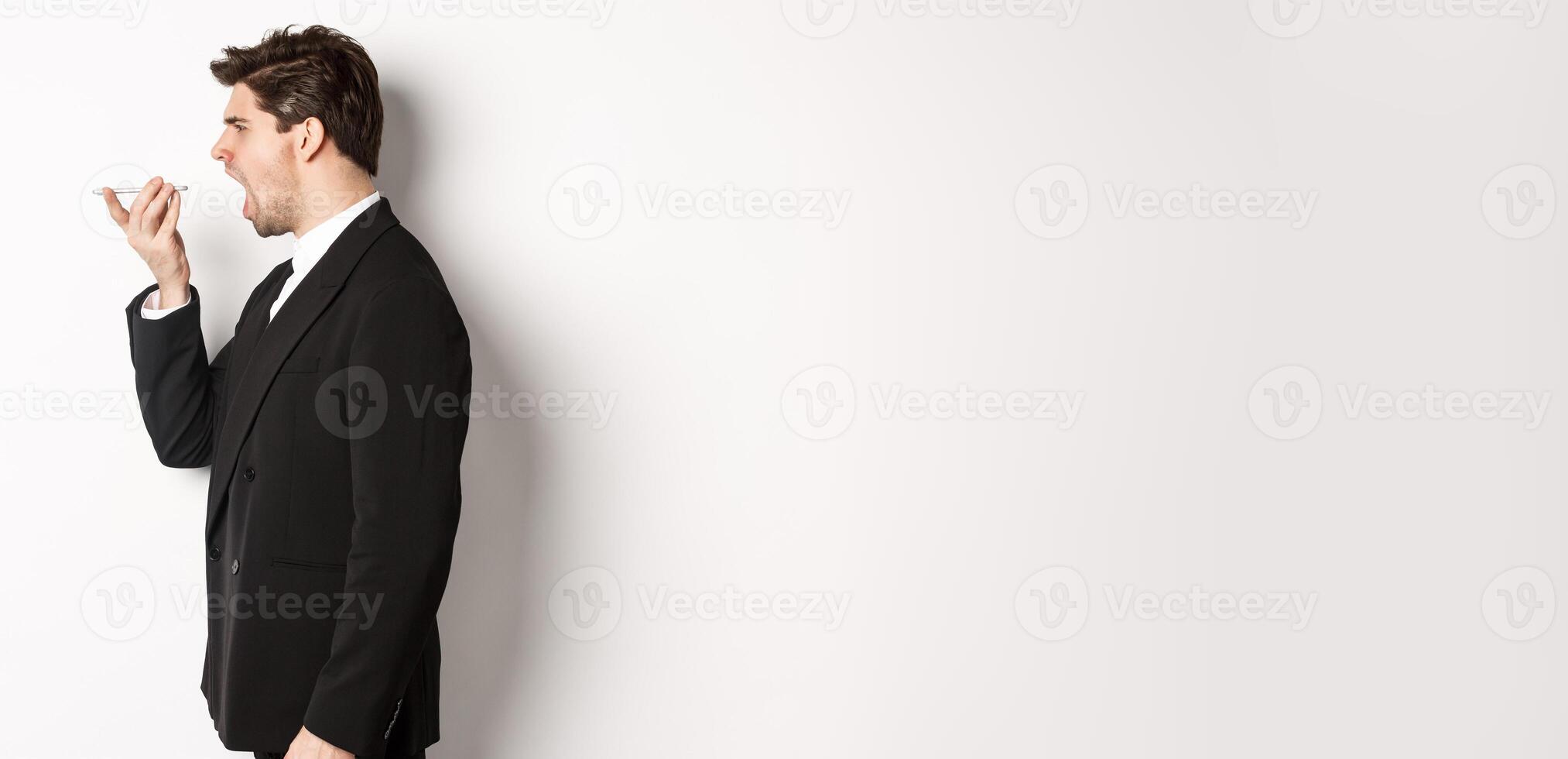 Profile shot of angry businessman in black suit, shouting at speakerphone and looking mad, recording voice message, standing over white background photo