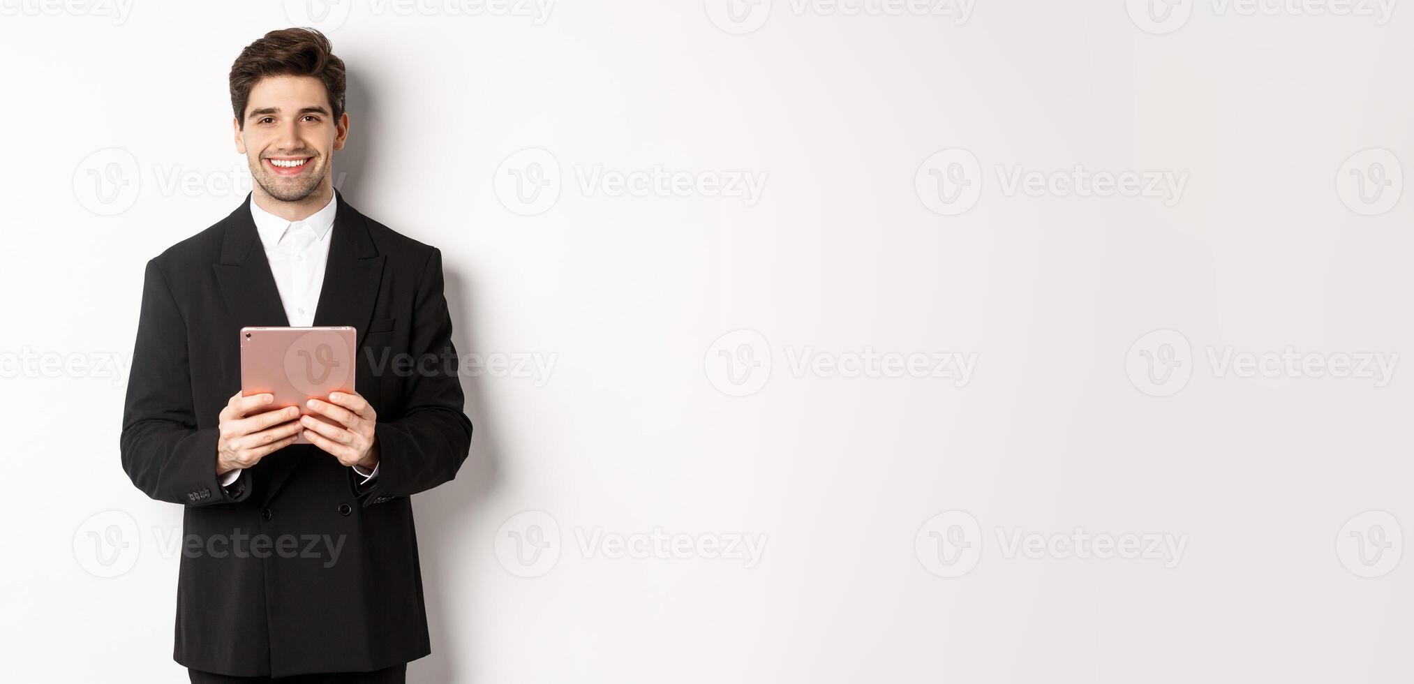 Image of handsome businessman in trendy suit, holding digital tablet and smiling, standing against white background photo