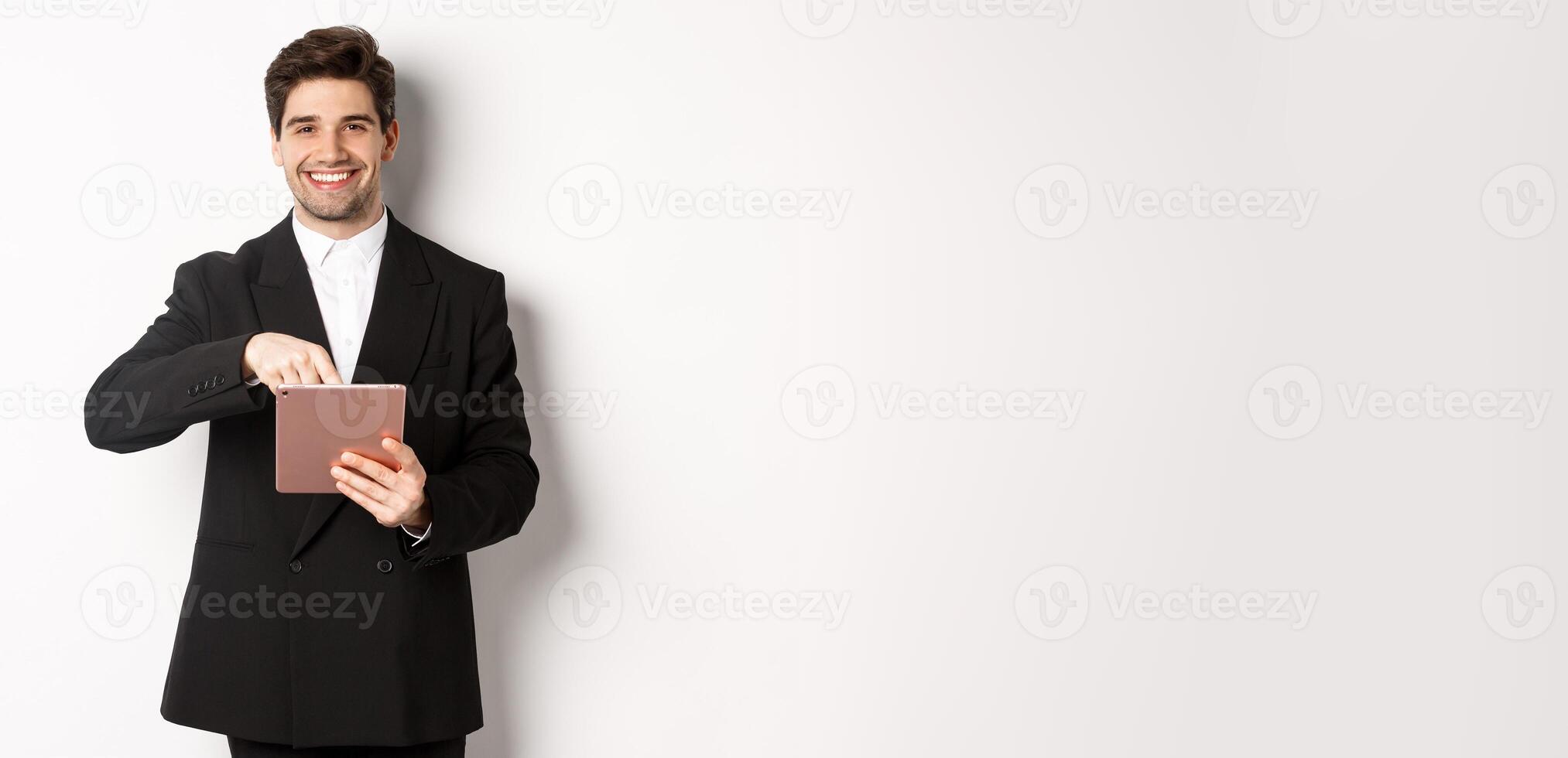 Portrait of handsome, stylish male entrepreneur in black suit pointing at digital tablet, showing something online, standing against white background photo