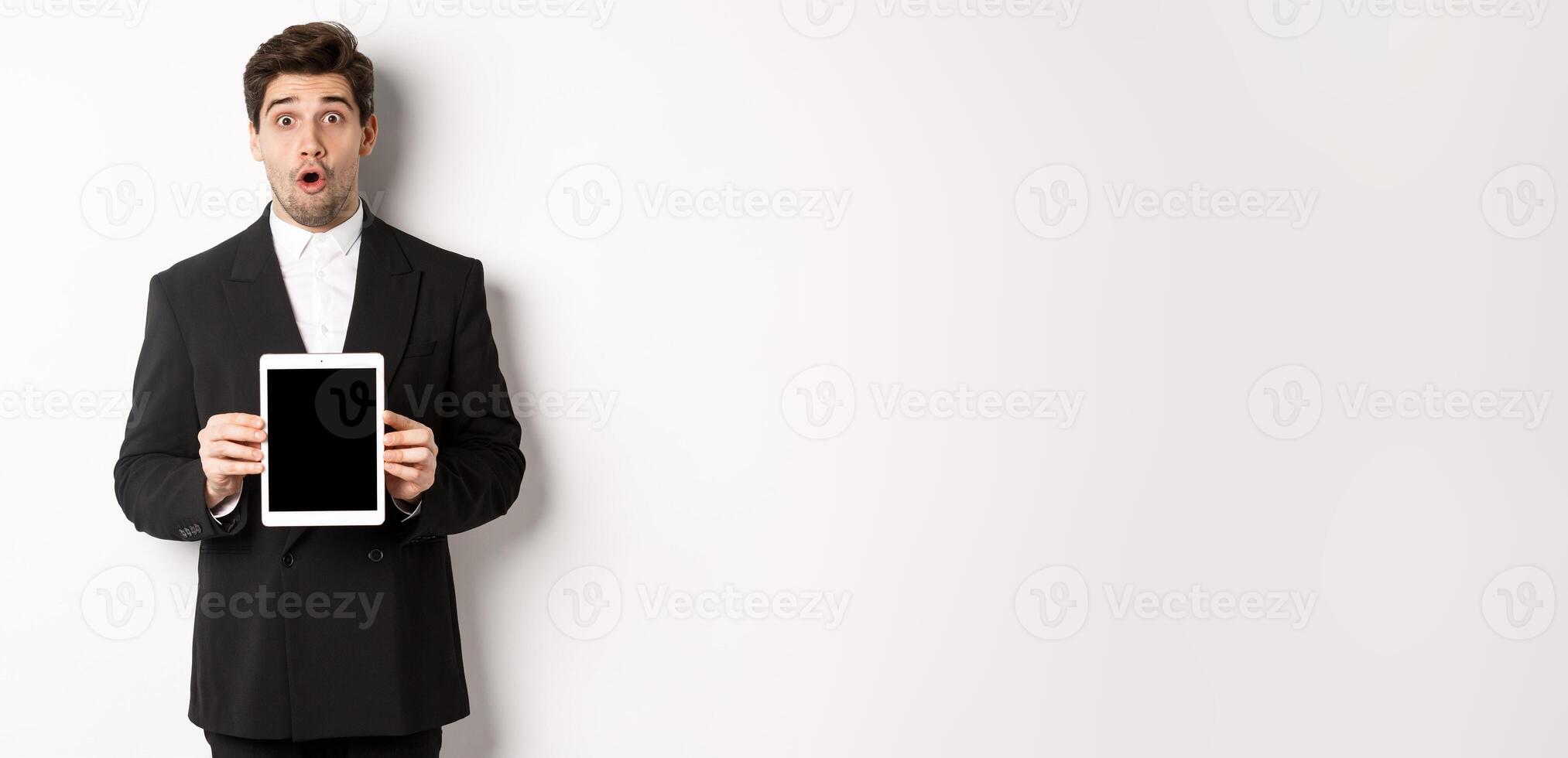 Image of surprised handsome man in black suit, showing digital tablet screen and looking amazed, standing against white background photo