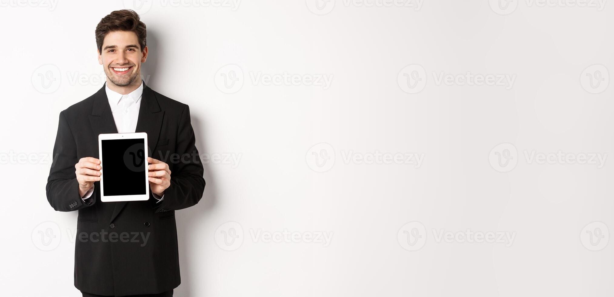 Portrait of handsome businessman in trendy suit, showing digital tablet screen and smiling, standing against white background photo