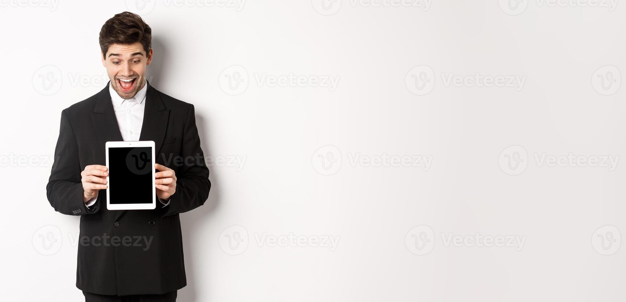 Portrait of amazed handsome man in black suit, showing digital tablet screen with excitement, standing over white background photo
