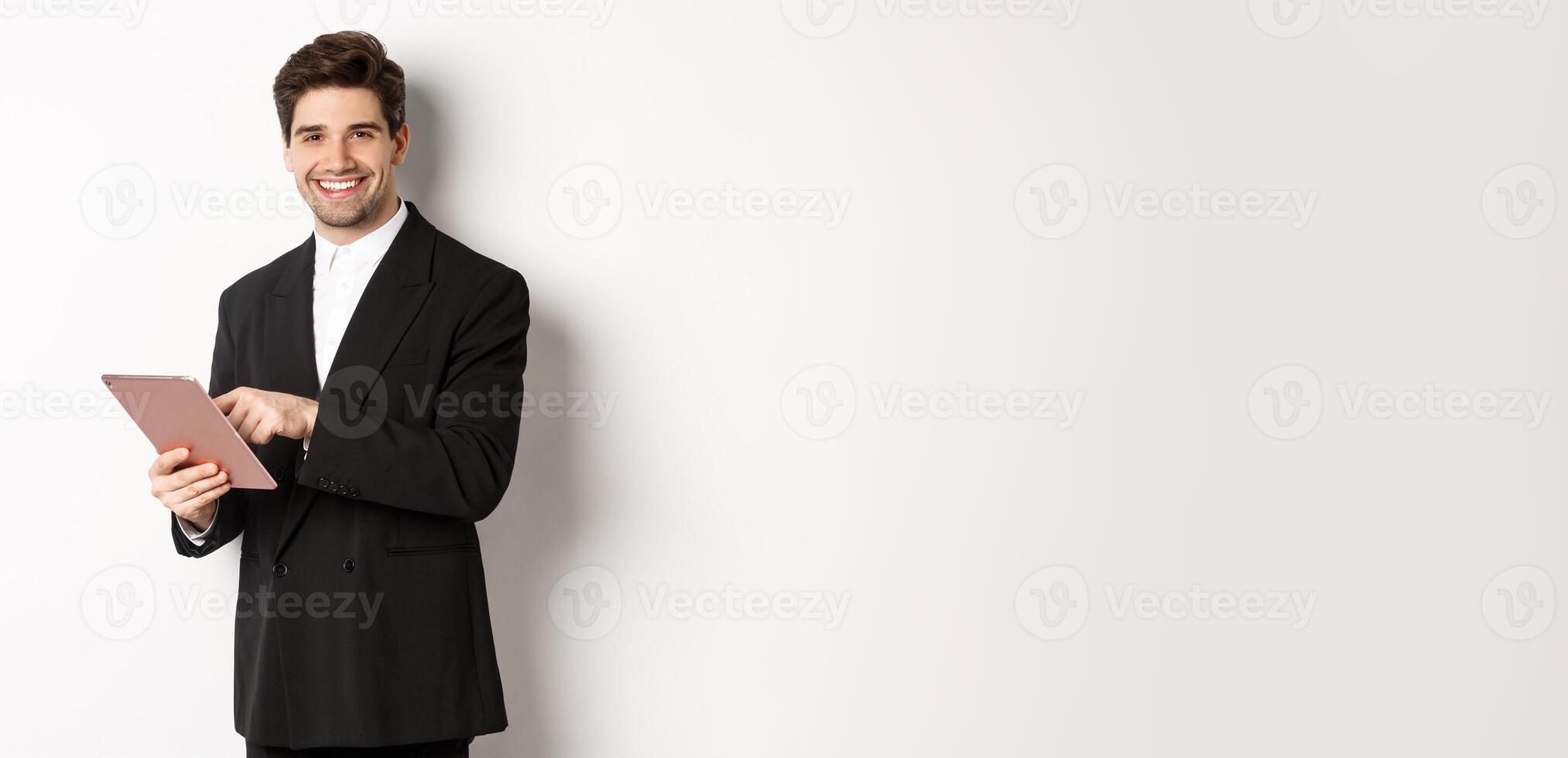 Image of handsome businessman in black suit, using digital tablet and smiling, standing against white background photo