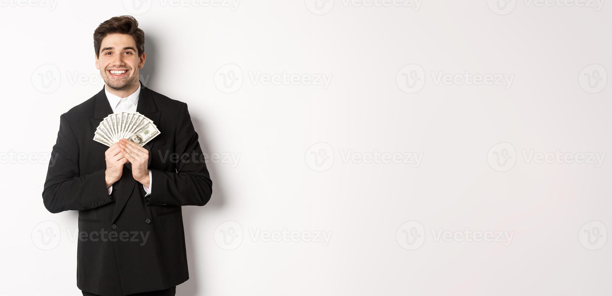 Portrait of happy and pleased handsome man in suit, hugging money and looking satisfied, standing over white background photo