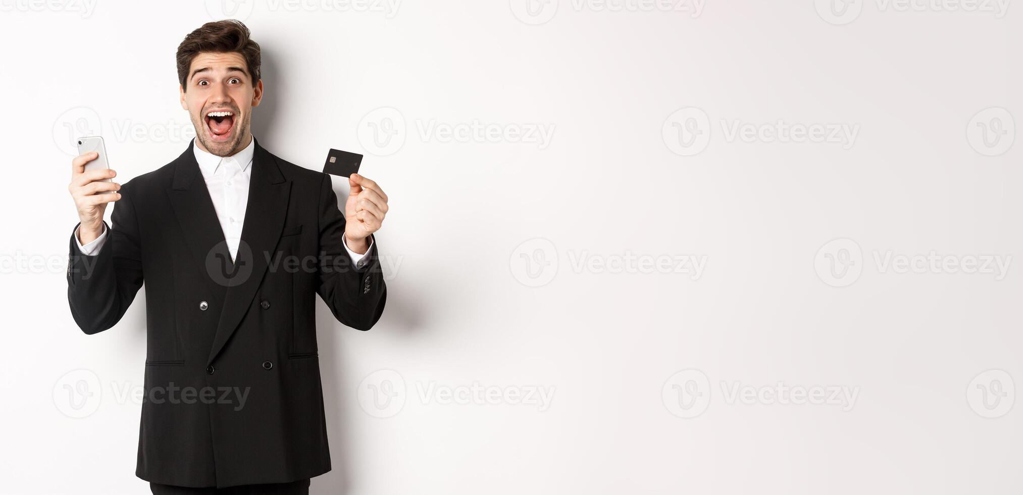 Handsome businessman in black suit smiling, showing credit card and money, looking amazed, standing against white background photo